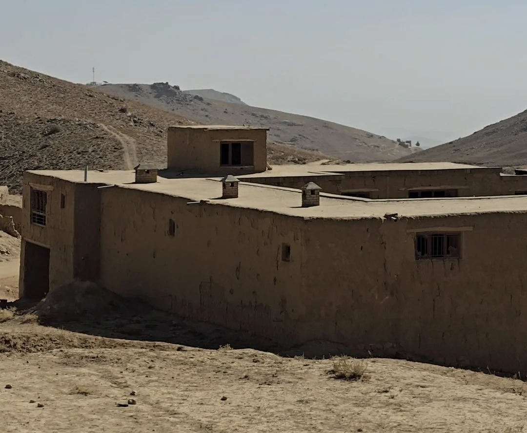A desert landscape with a large adobe-style building in the foreground and hills in the background.