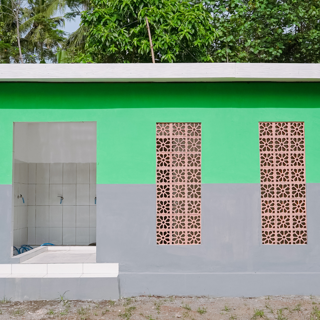 A partially constructed building with a green upper wall, a gray lower wall, and two sections of decorative pink bricks. An open doorway reveals a tiled interior with pipes, and there is a small step at the entrance. Trees are visible above the roof.
