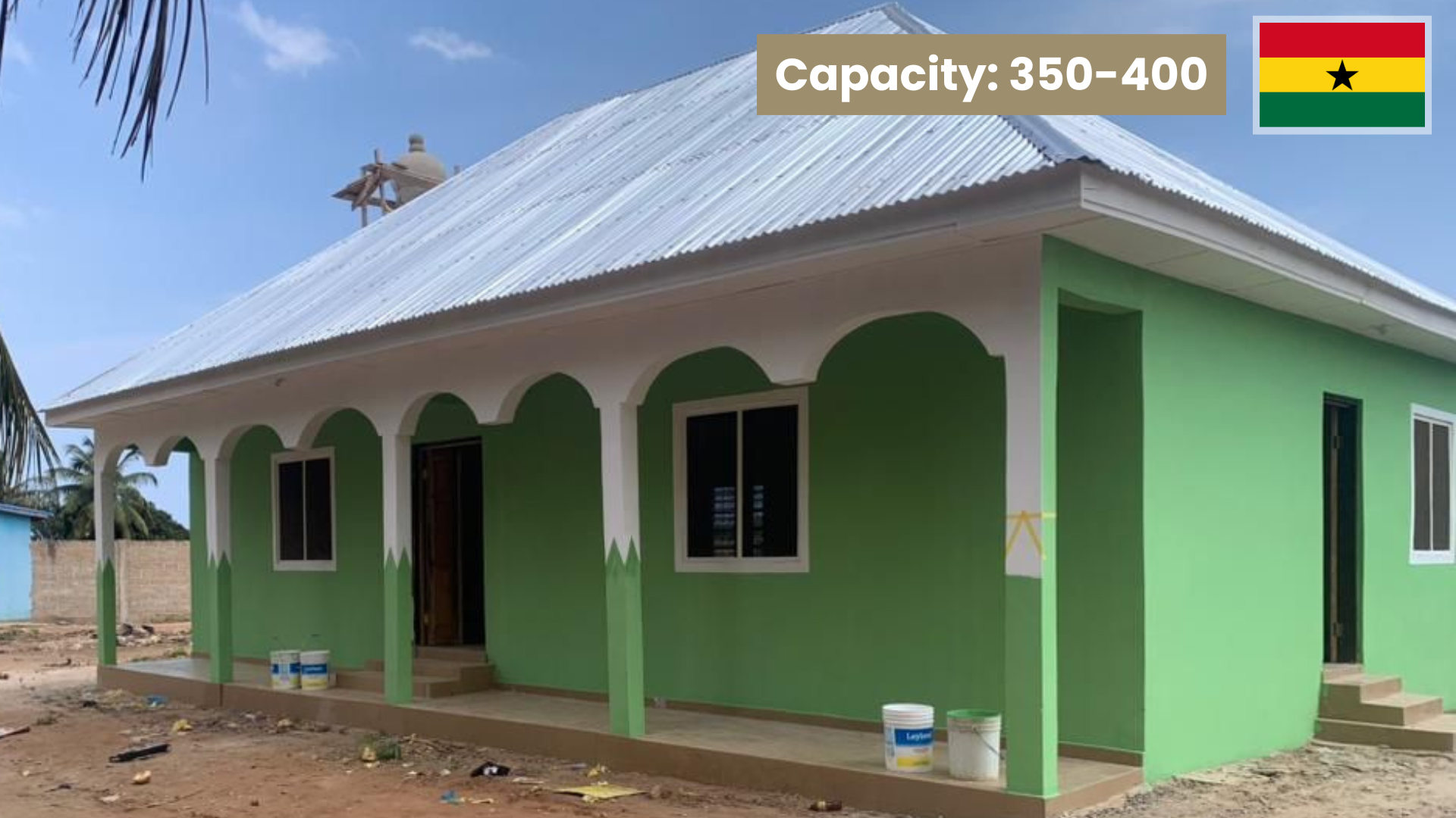 Bright green house with a metal roof, decorative arches on the porch, and several paint buckets in front, located in Ghana.