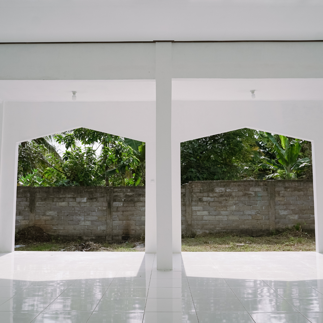 View of a modern open porch or patio with white tiled floor, white walls, and open archways revealing a brick wall with green foliage and trees in the background.