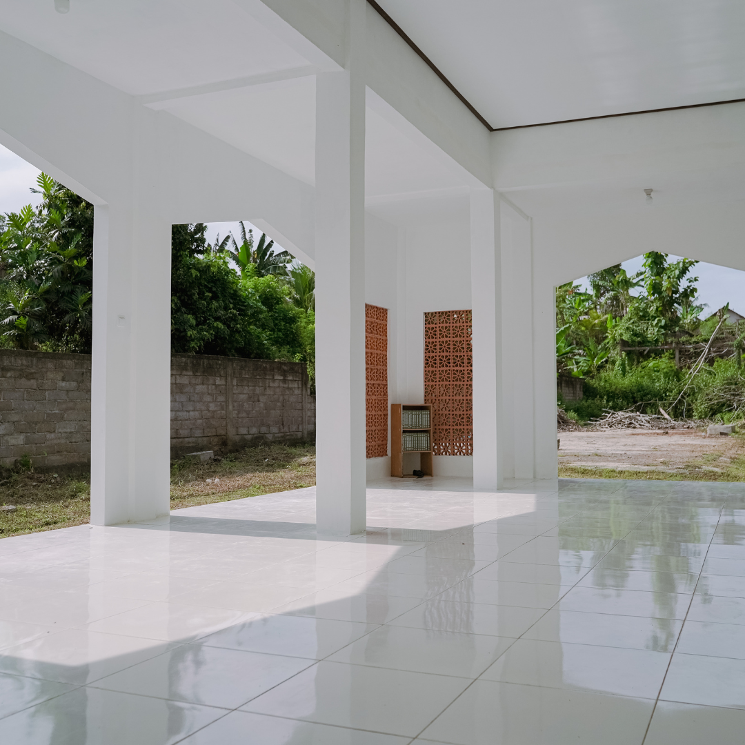 Empty white outdoor patio with tile floor, supported by beams, and trees and a brick wall in the background.