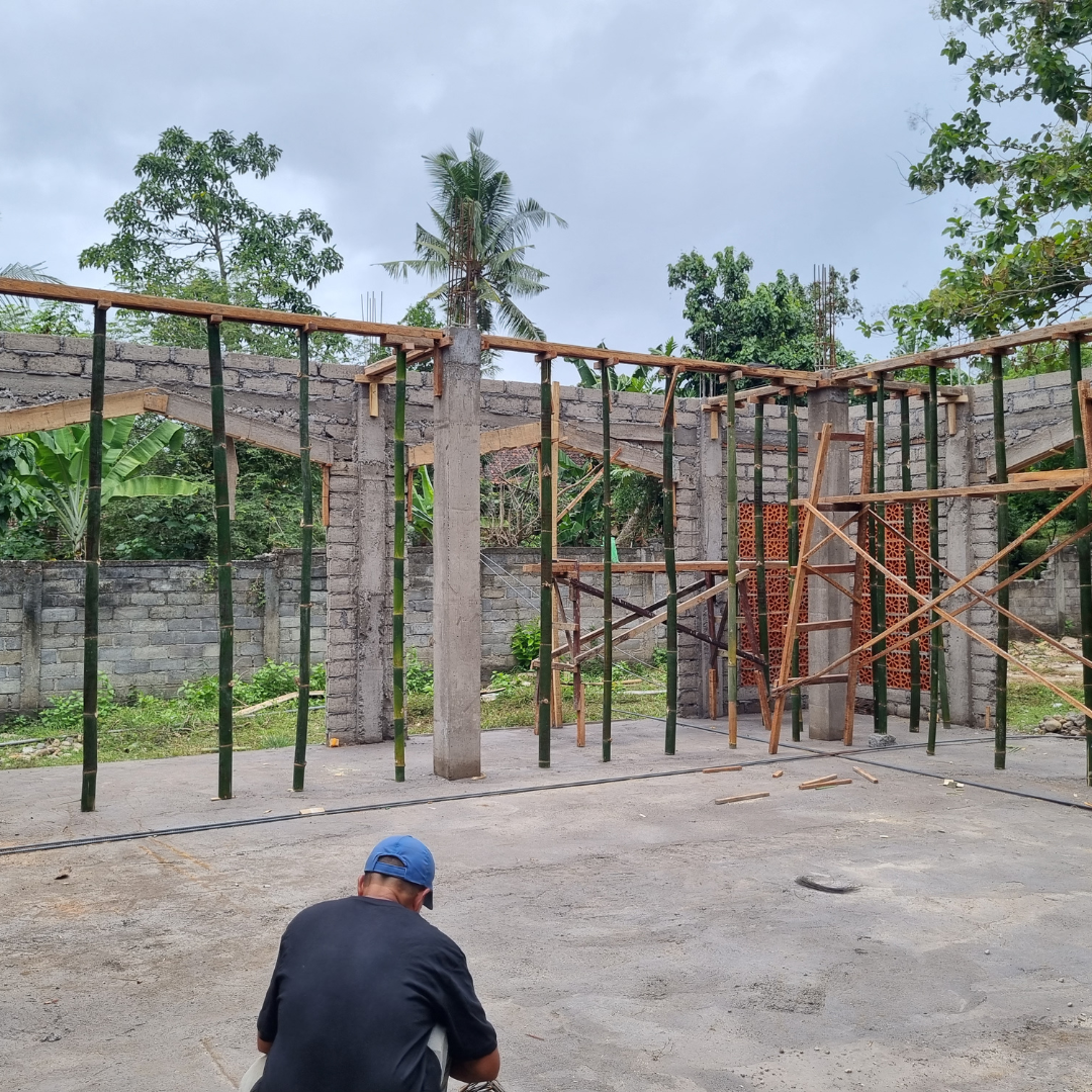 Construction site with concrete columns and unfinished brick walls, scaffolding around the structure, and a man kneeling on the ground wearing a black shirt and blue cap.