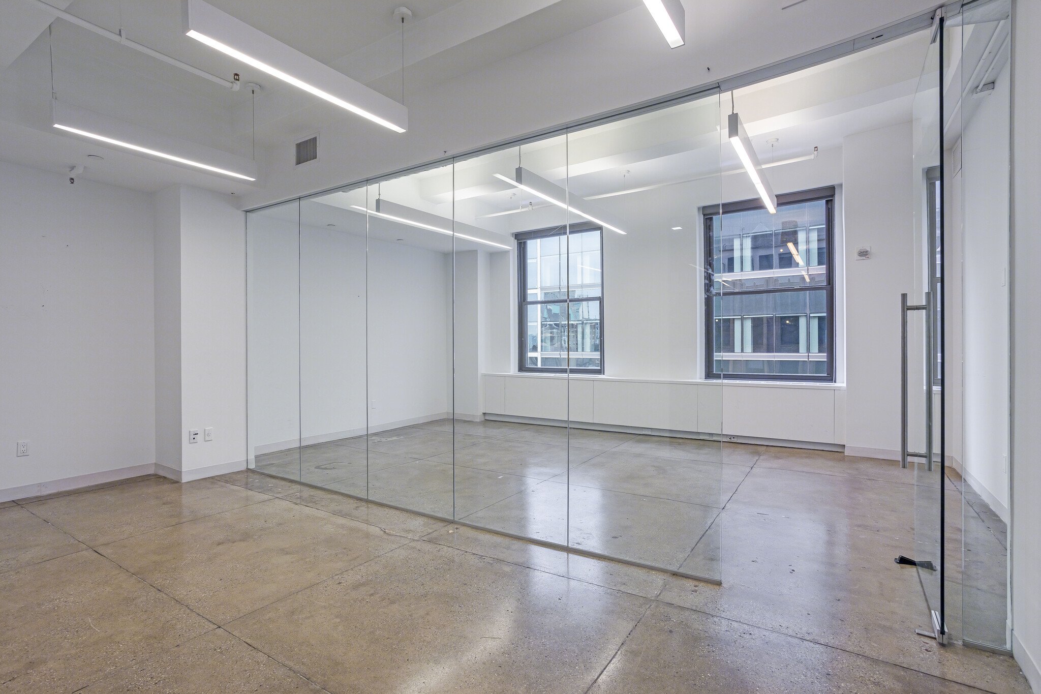 Empty office space with concrete floors, white walls, large windows, glass wall and door, and modern ceiling lighting.