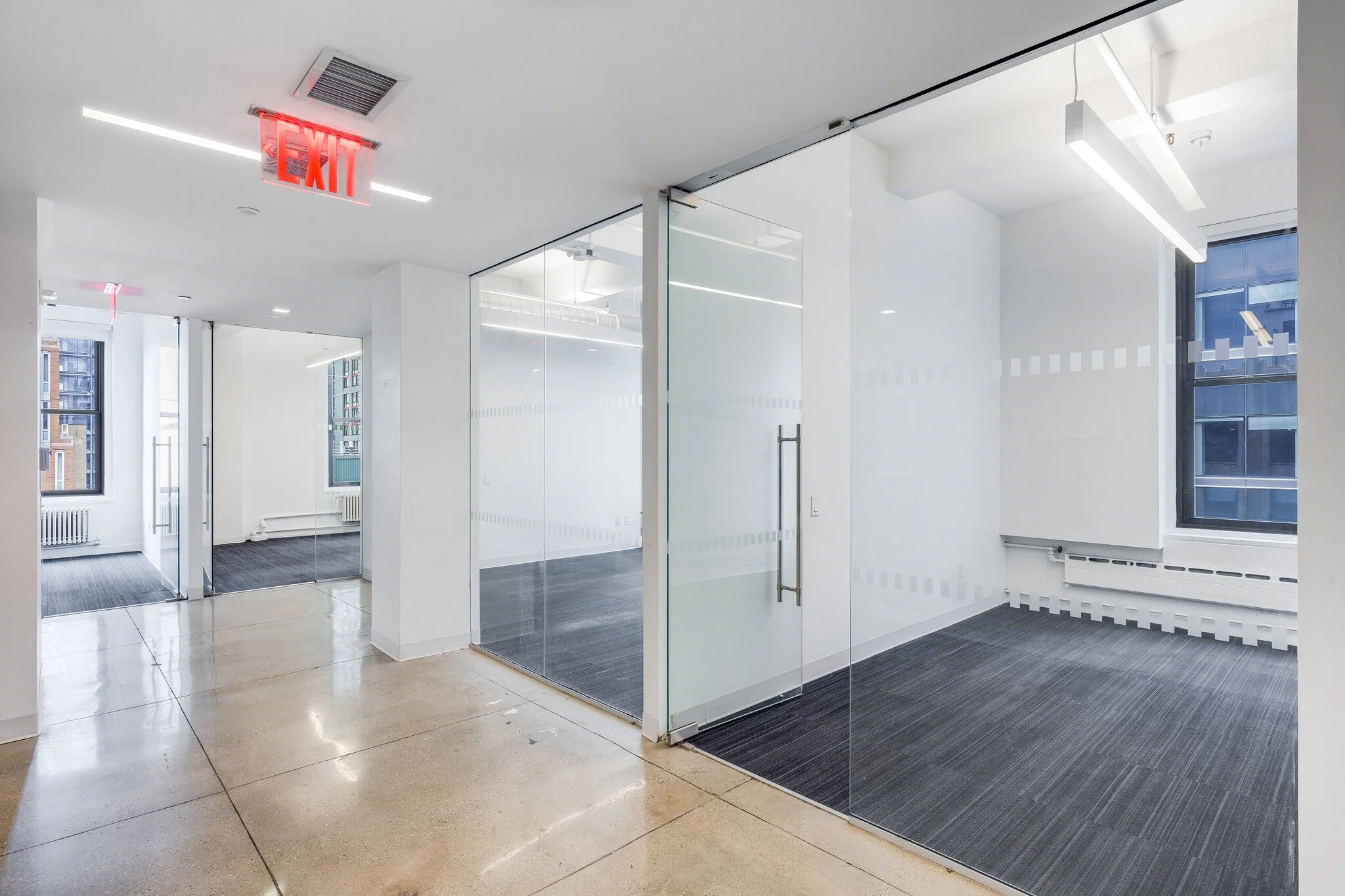 Empty office space with glass walls, large windows, and an EXIT sign on the ceiling.