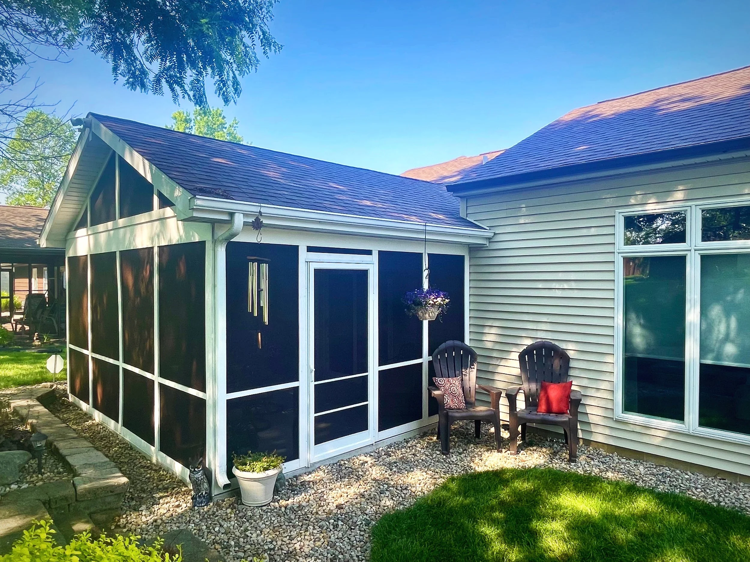 Backyard patio with two brown chairs, red and patterned pillows, a hanging flower basket, and a screened-in porch attached to a house with beige siding and a large window. Gravel ground and green grass are visible along with shaded trees.