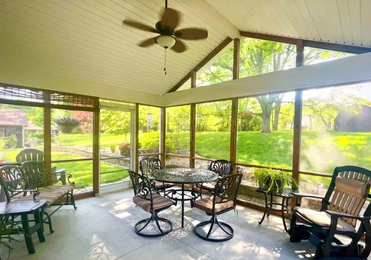 Sunroom with screened windows, wooden ceiling, ceiling fan, metal dining table, six chairs with striped cushions, potted plant, and view of green yard with trees and grass