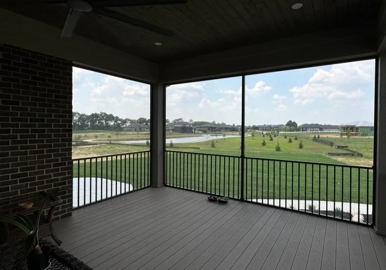 View from a screened porch overlooking a green lawn, trees, and houses under a partly cloudy sky.