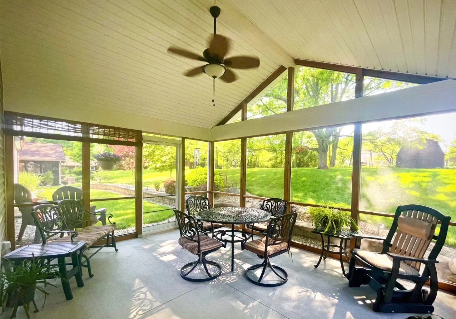 Screened porch with outdoor furniture, including a round metal table with five chairs, two cushioned armchairs, a rocking chair, and potted plants, overlooking a green yard with trees and a shed.