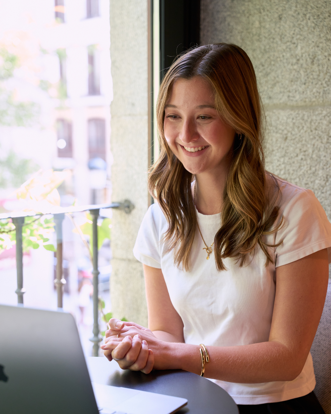 A woman with shoulder-length brown hair smiling while sitting at a table near a window, using a laptop.
