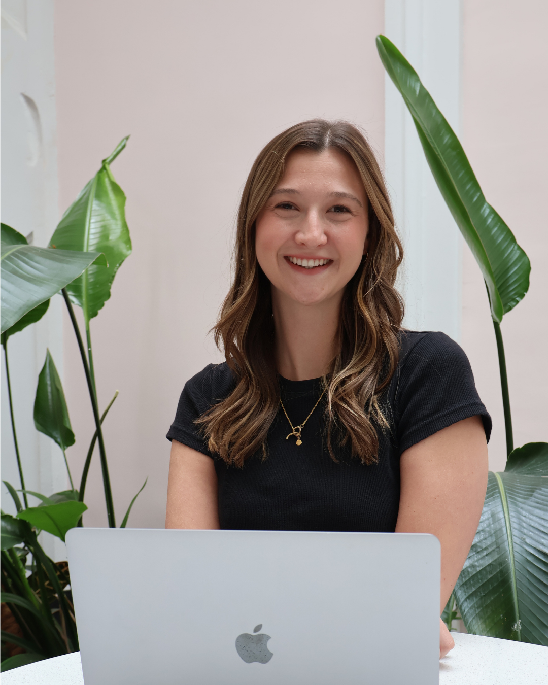 A woman with wavy brown hair, smiling, sitting at a desk with a laptop, surrounded by large green plants.