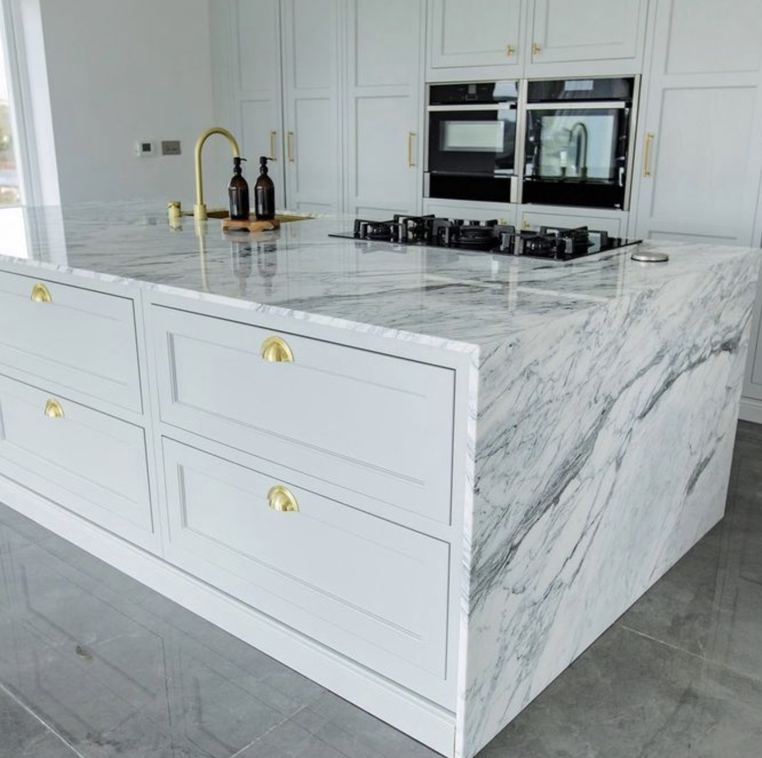 A kitchen island with a white marble countertop and matching sides, featuring gold handles. In the background, there are built-in white cabinets, a stove, a microwave, and a sink with a gold faucet. Three brown bottles are on the island.