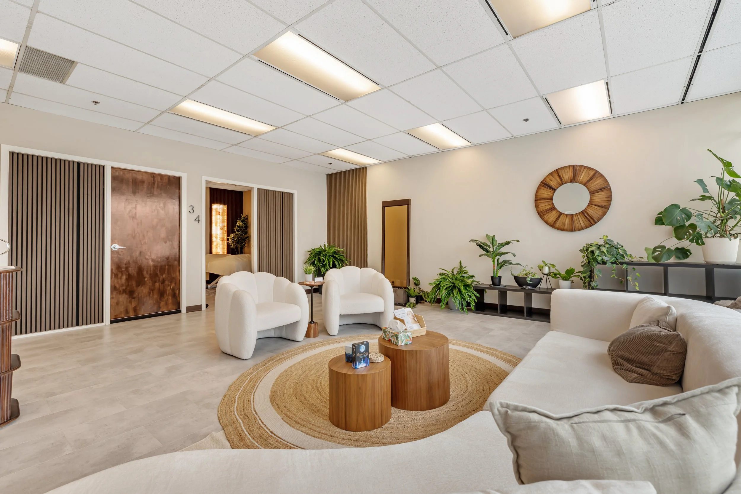 Modern living room with white curved sofa, matching armchair, round wooden coffee tables, beige rug, large potted plant, built-in shelving, and decorative mirror. Showing the treatment rooms with ambient lighting.
