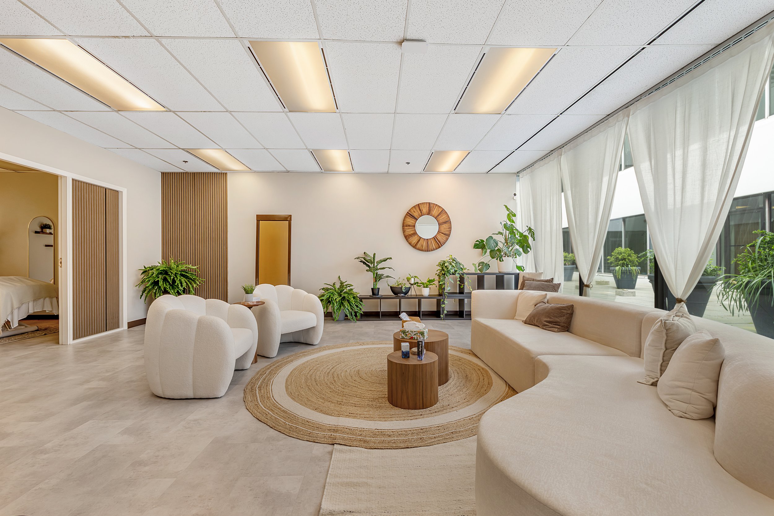 Modern minimalist client lounge area  with white curved sofa, beige throw pillows, wooden coffee tables, potted plants, and a black metal and wood shelf against a white wall. Large window with white curtains and ceiling with recessed lighting.
