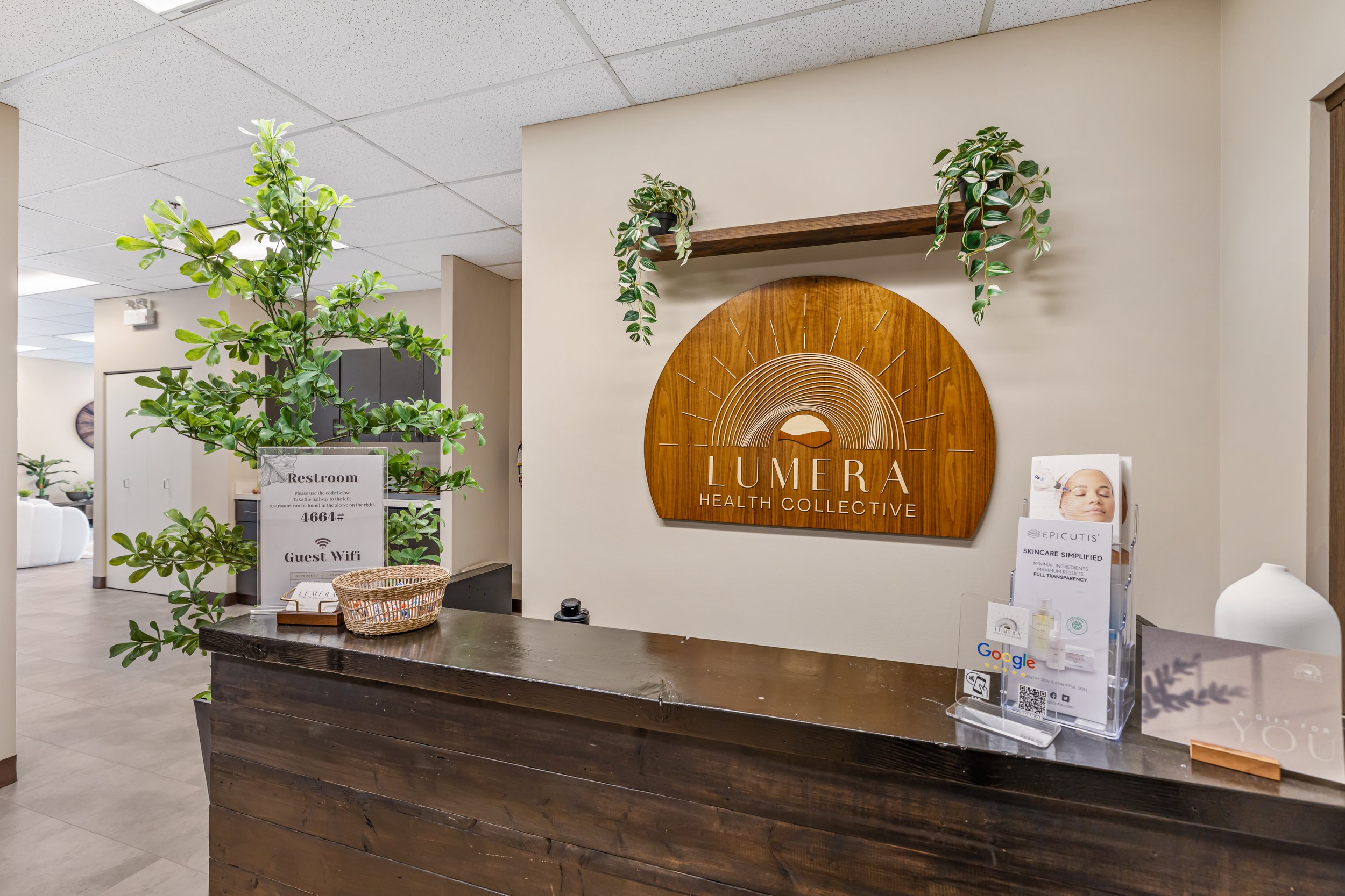 Reception area with wooden desk, potted tree, and wall decor featuring a sunrise and the words "LUMERA HEALTH COLLECTIVE."