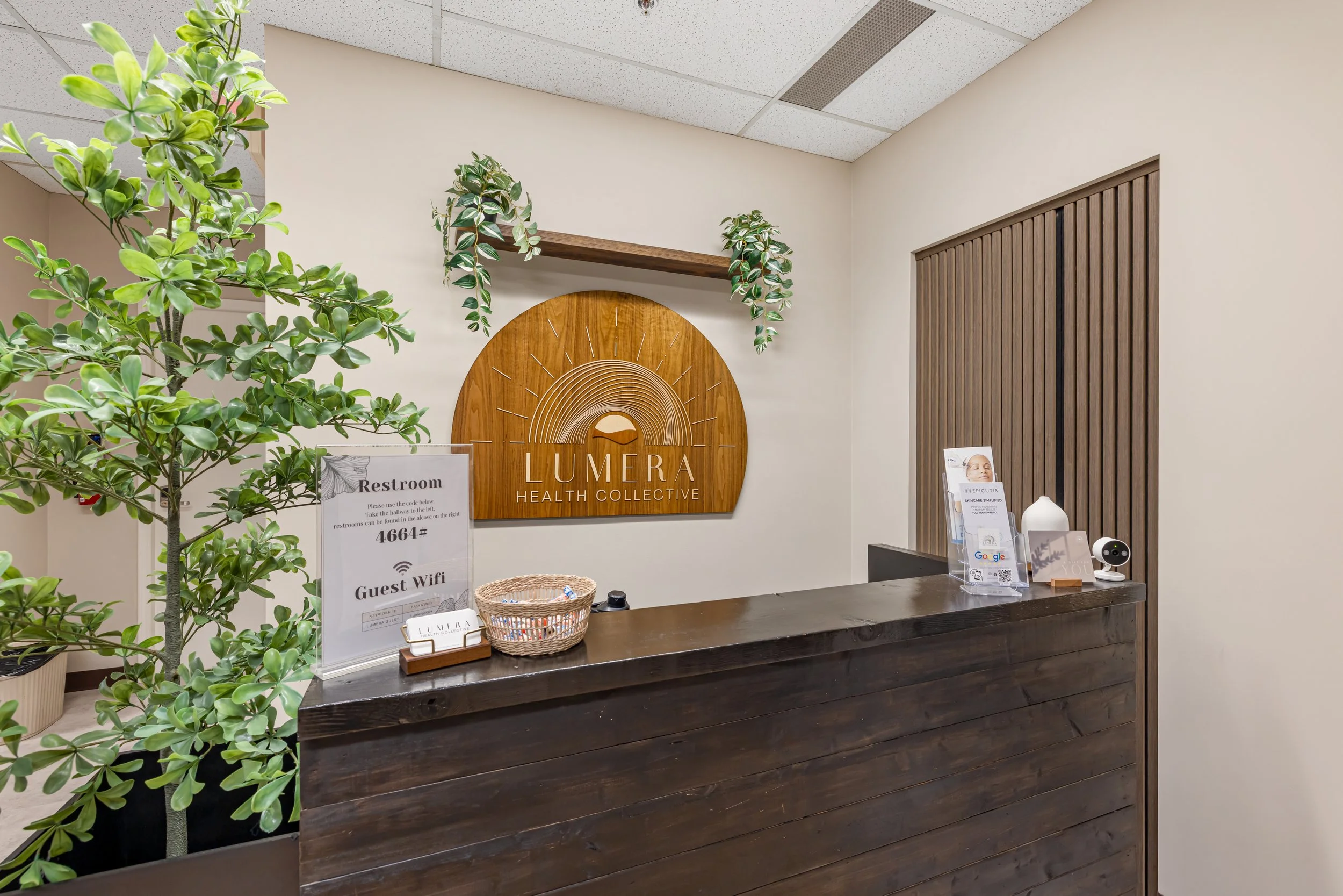 Reception area with wooden desk, potted tree, and wall decor featuring a sunrise and the words "LUMERA HEALTH COLLECTIVE."