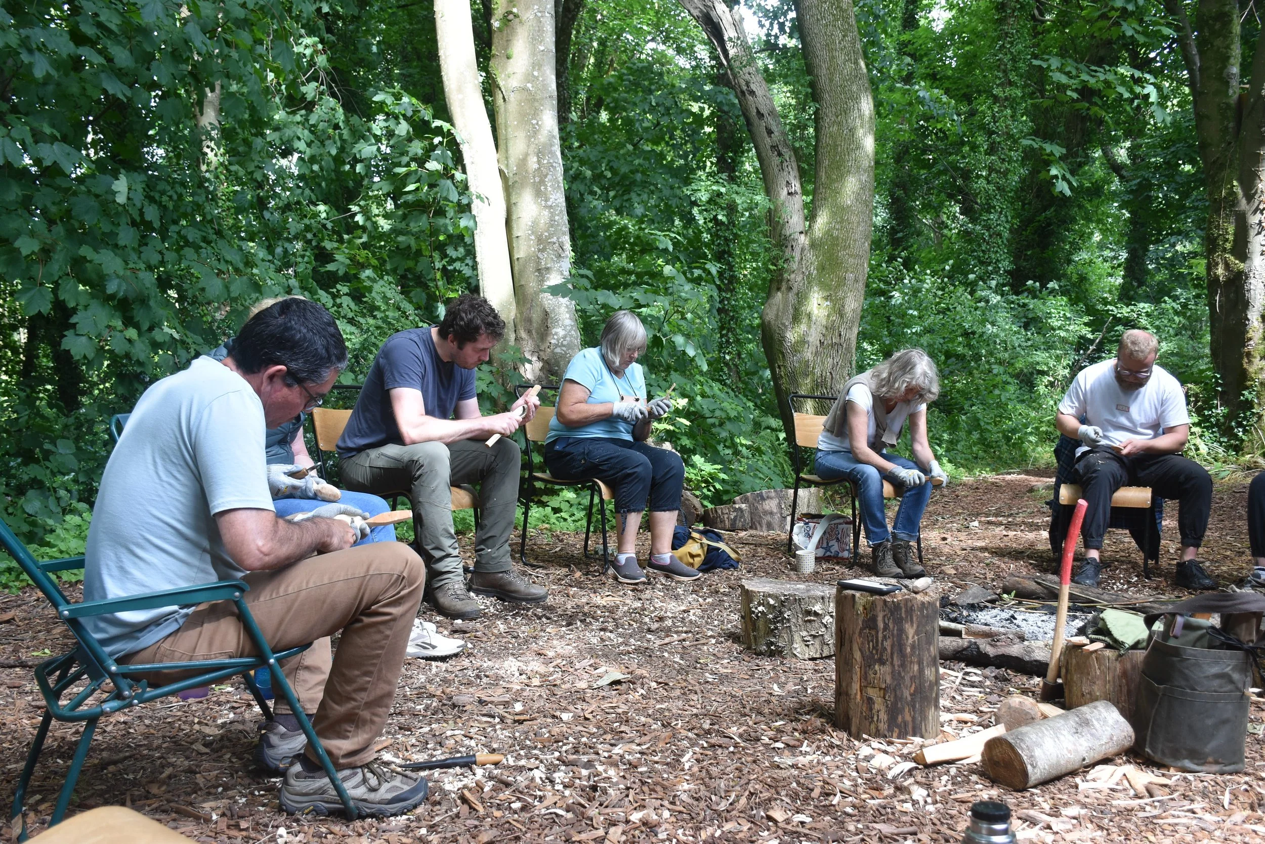 A group of six people sitting on chairs in a wooded outdoor area, examining and working on small objects in their hands, with logs and a campfire setup nearby.