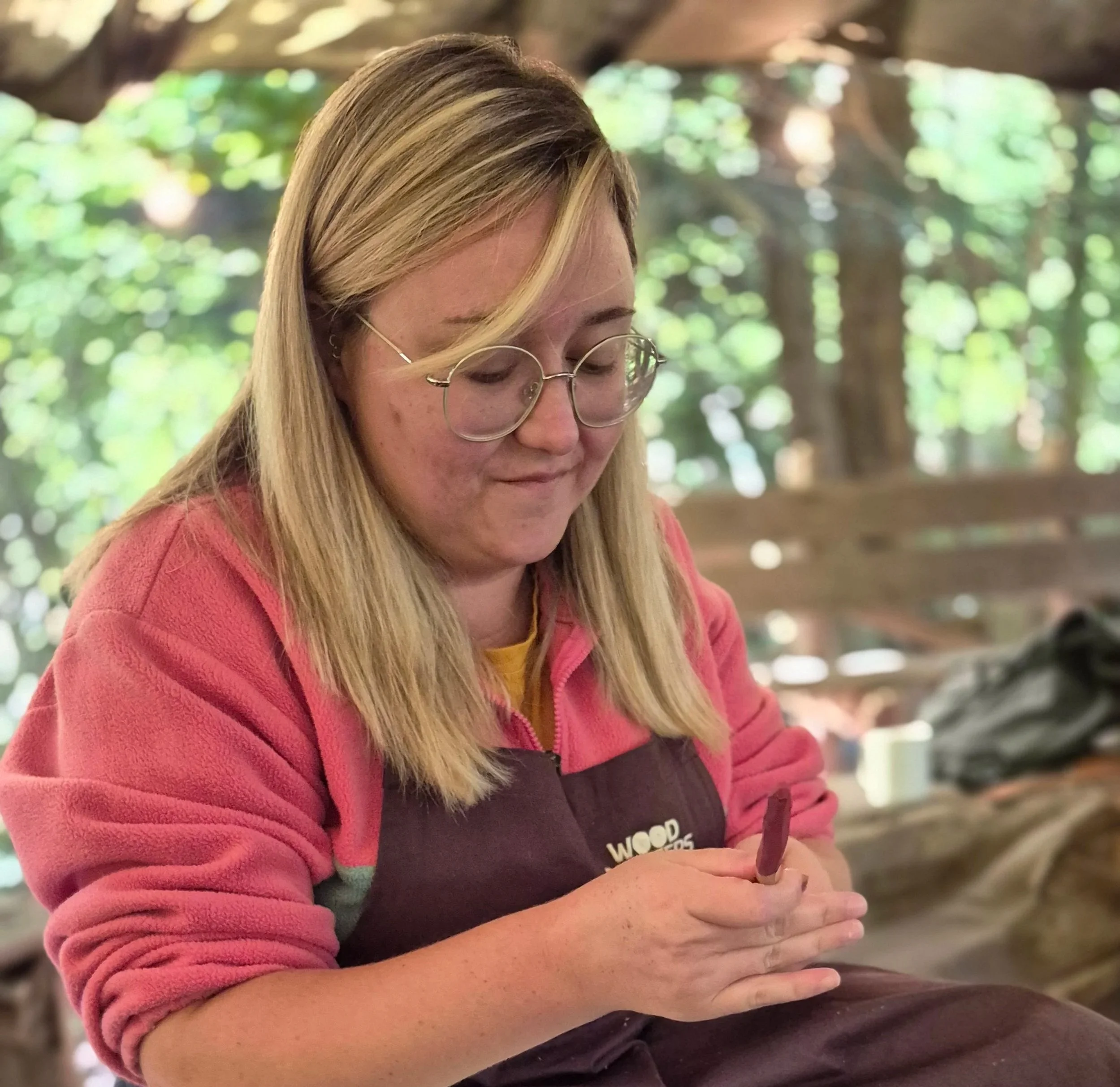 A young woman with blonde hair and glasses carving wood with a small chisel outdoors.
