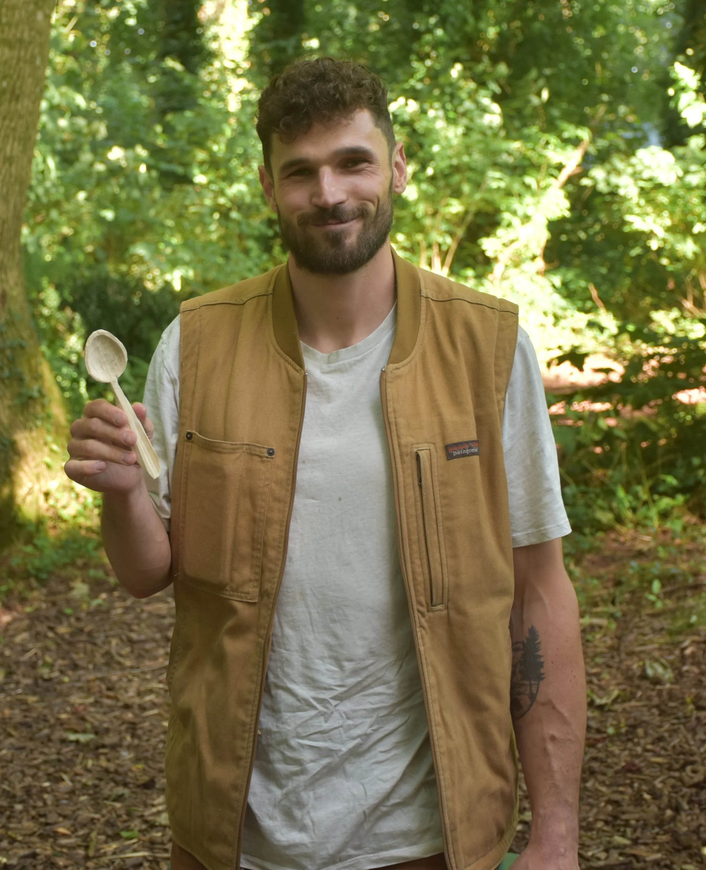 A smiling man with a beard and curly hair, wearing a tan vest over a white t-shirt, holding a small wooden spoon, standing in a wooded area with green foliage.