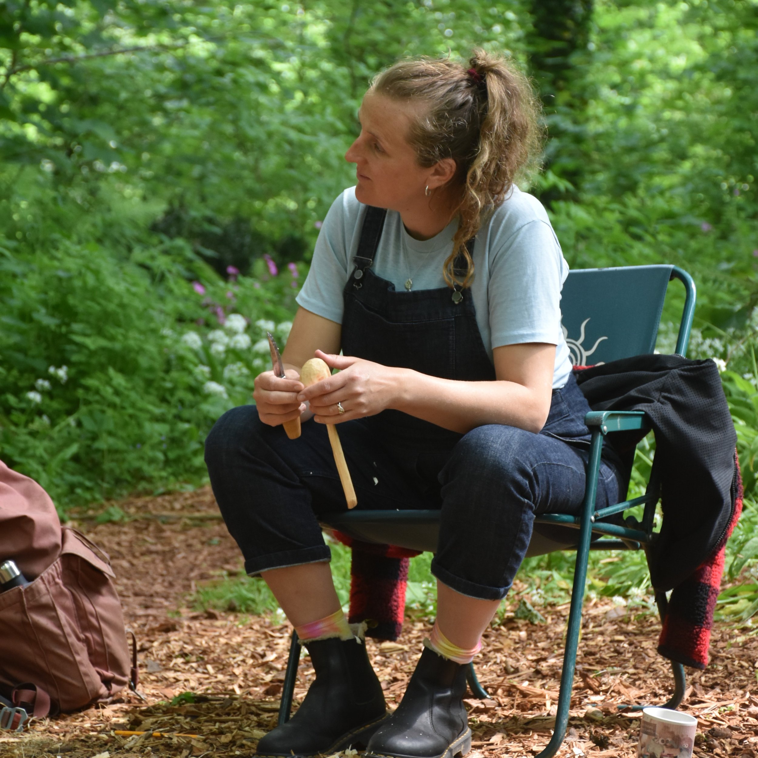 Woman sitting on a camping chair in a forest, holding a wooden mallet and a small round object, with a backpack on the ground nearby.