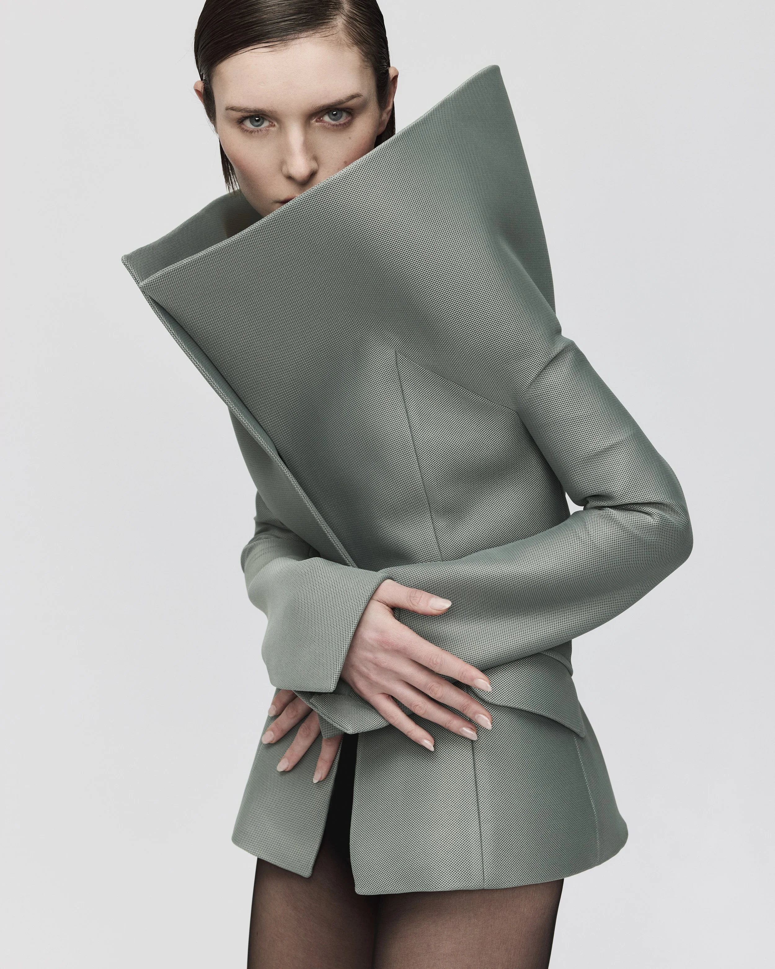 Frau in einem grünen Anzug hält einen großen, passenden grünen Kissen vor der Brust, blickt direkt in die Kamera, dunkles Haar, neutrale Hintergrund