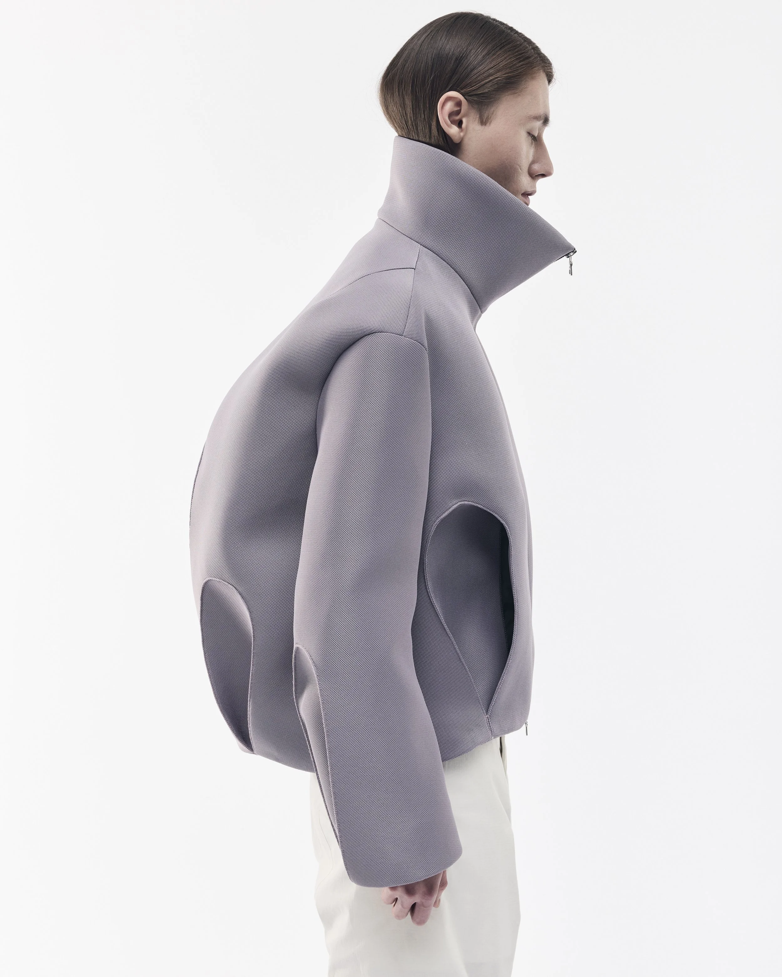 Ein Mann trägt eine moderne, graue, hoch geschlossene Jacke mit diskretem Schnitt und zipp am Kragen, fotografiert im Profil vor weißem Hintergrund.