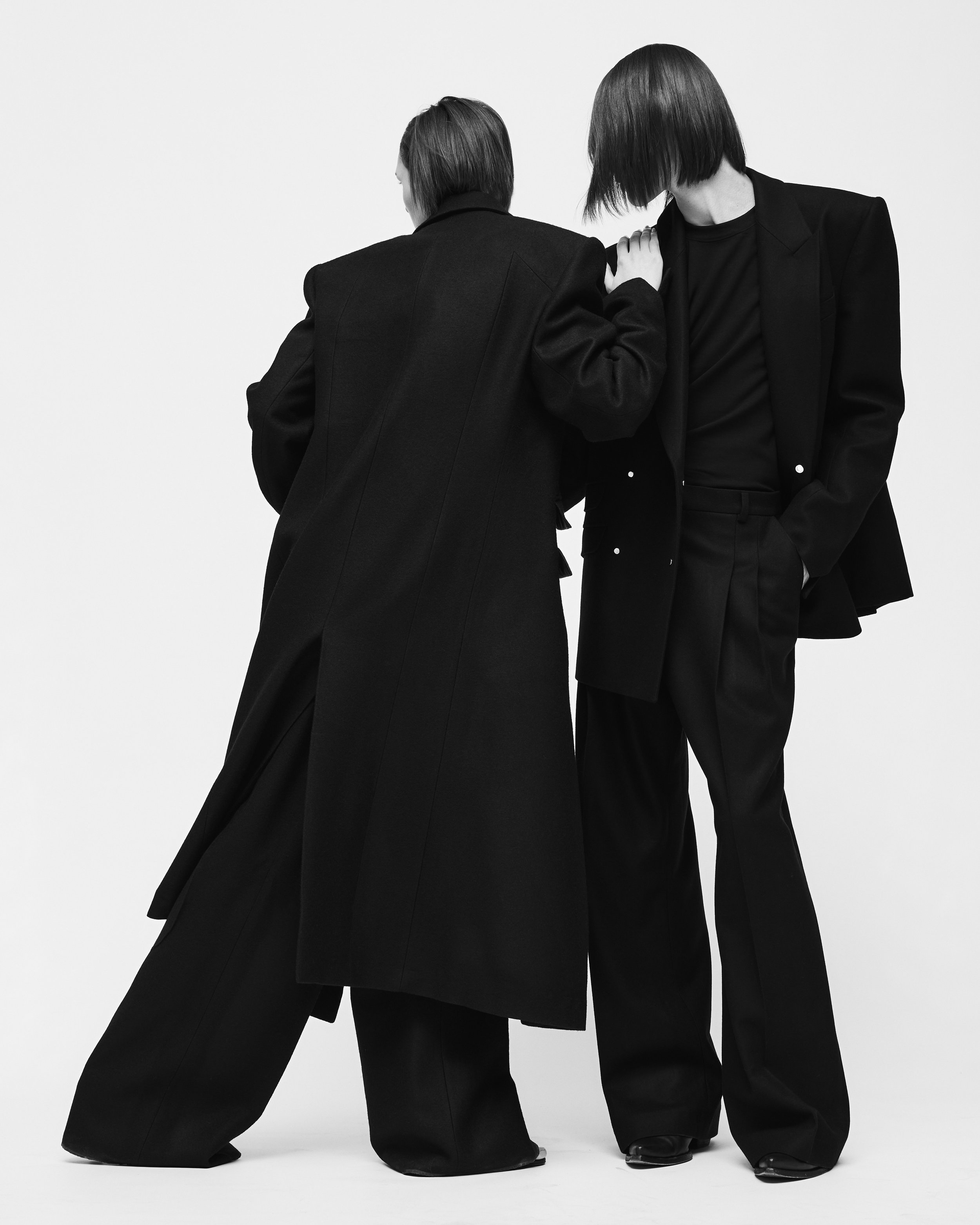 Zwei Menschen in schwarzen Anzügen stehen vor weißem Hintergrund, einer berührt die Schulter des anderen.