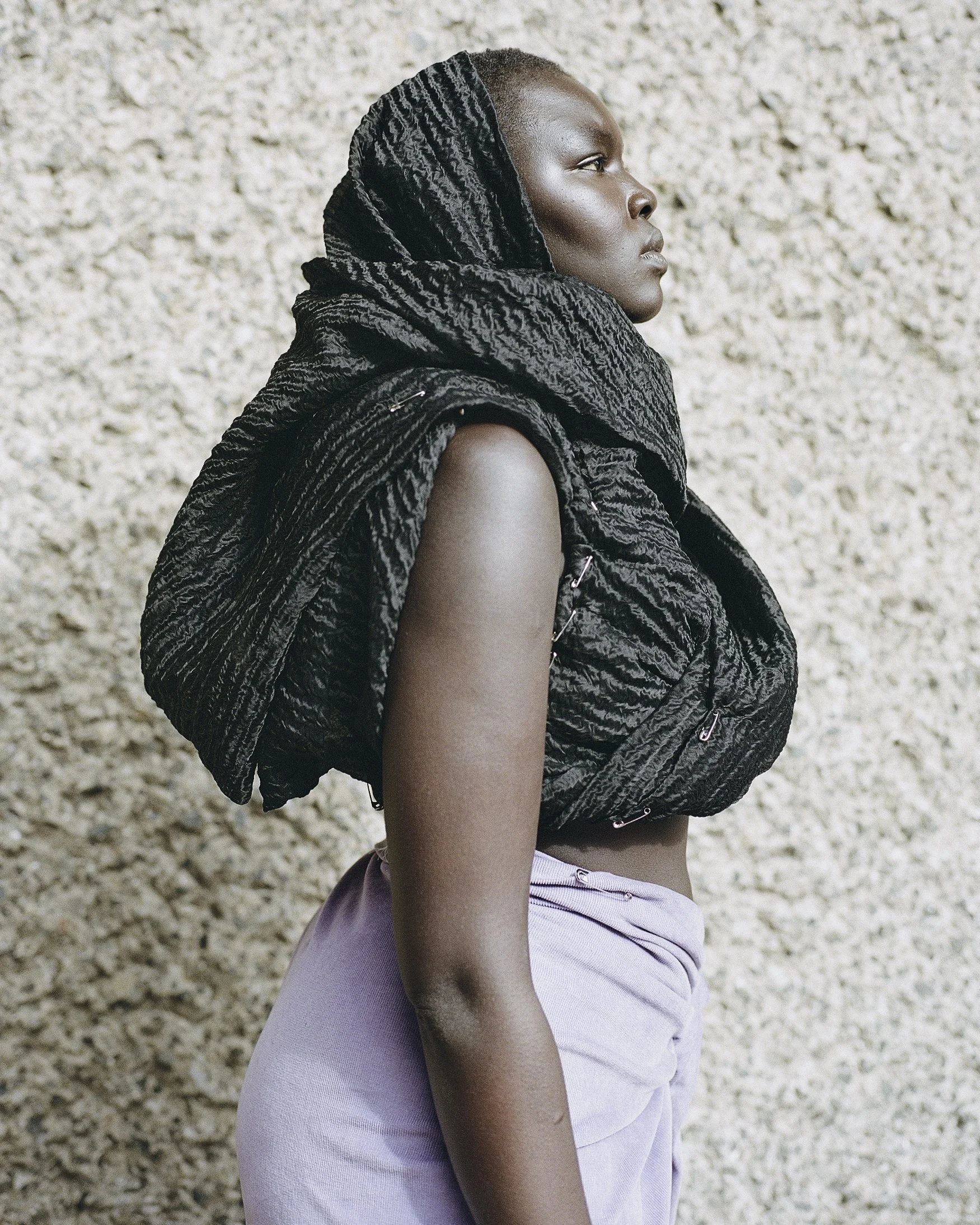 Frau mit schwarzem, gewickeltem Schal auf dem Kopf und schwarzer Weste vor einer beige Texturwand.