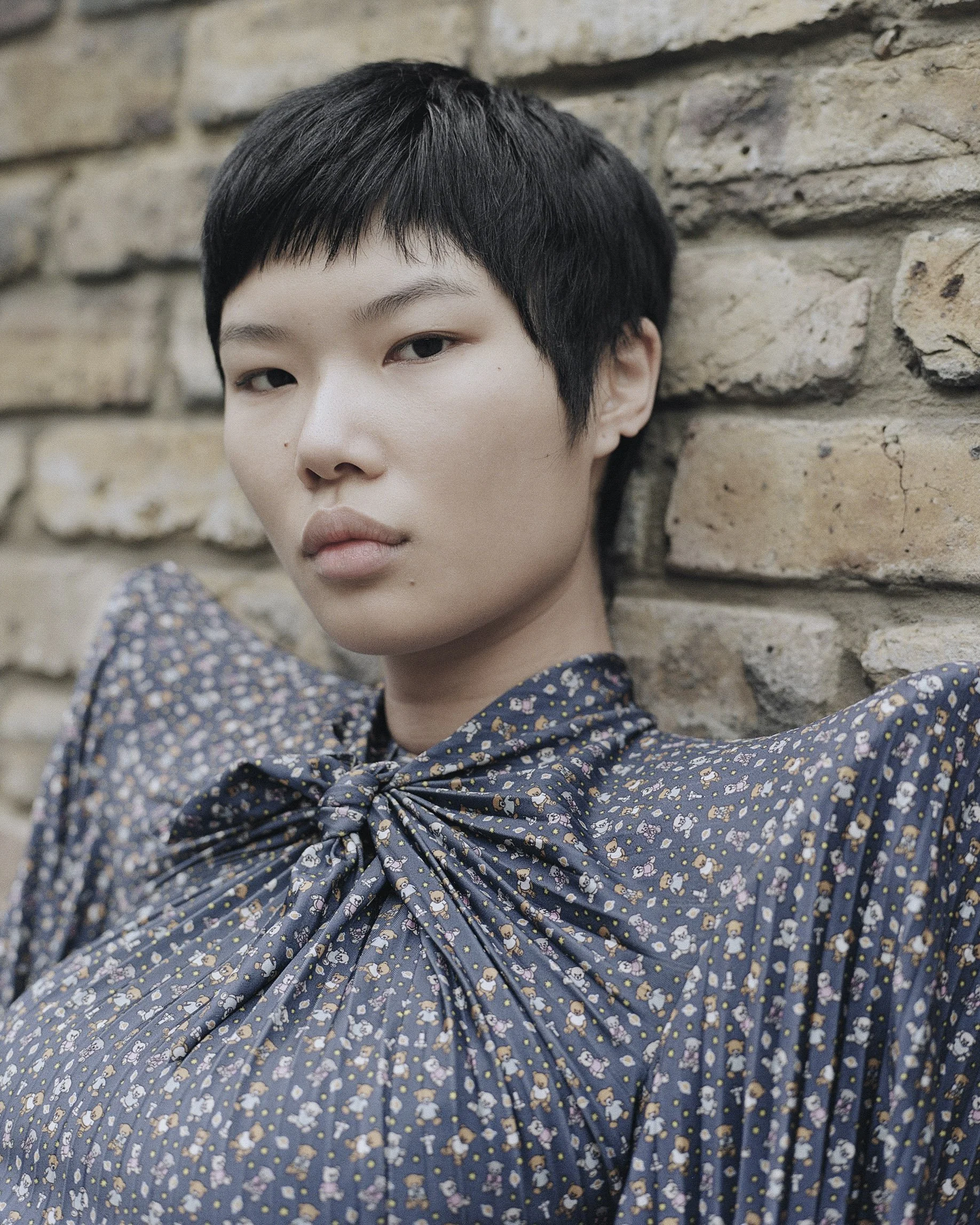 Ein junger Mensch mit kurzen schwarzen Haaren lehnt an einer Ziegelsteinmauer. Sie tragen ein dunkelblaues Kleid mit winzigen Blumenmustern und einem Schleifen-Kragen.