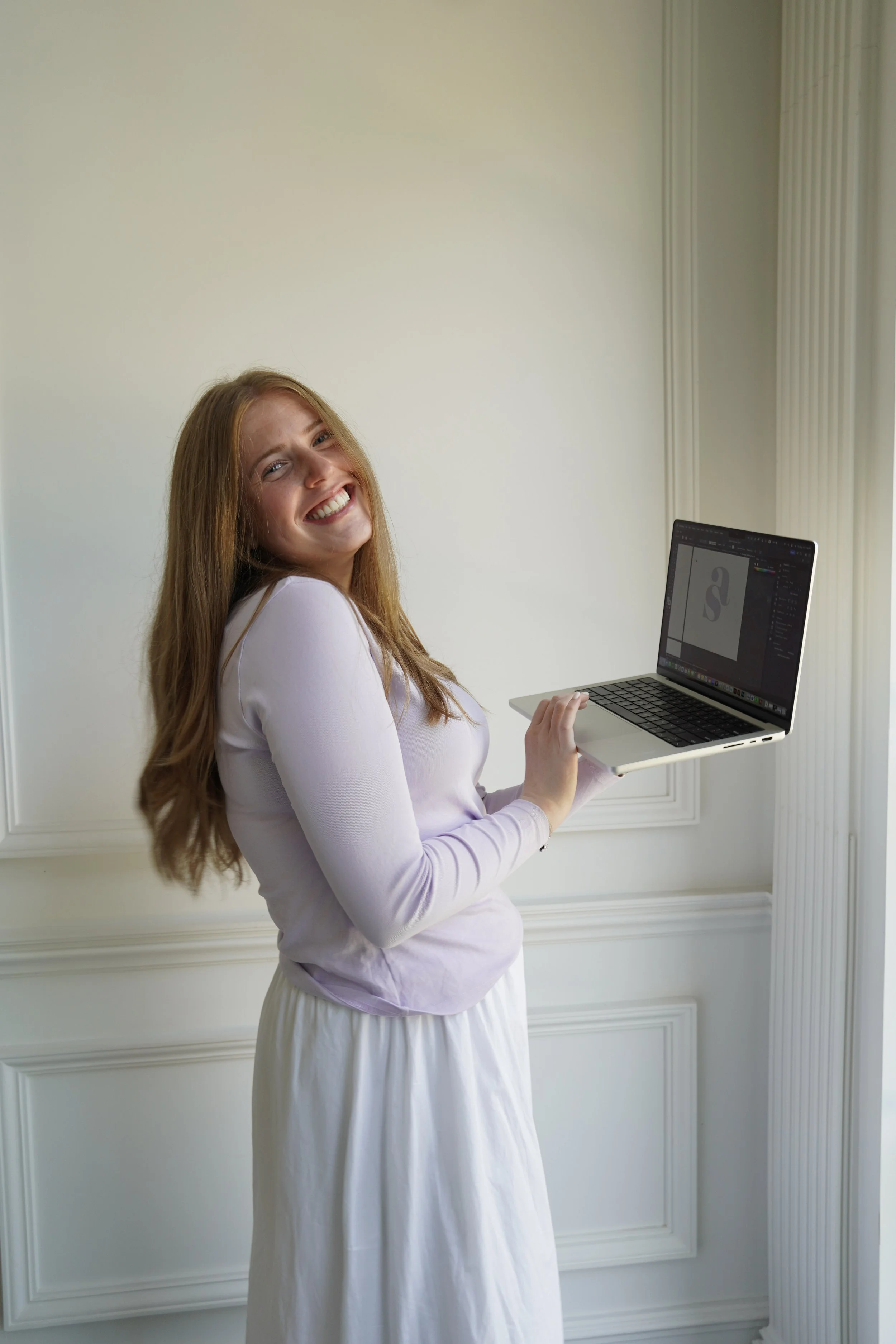 Smiling woman with long red hair holding a laptop in front of a white wall and window.
