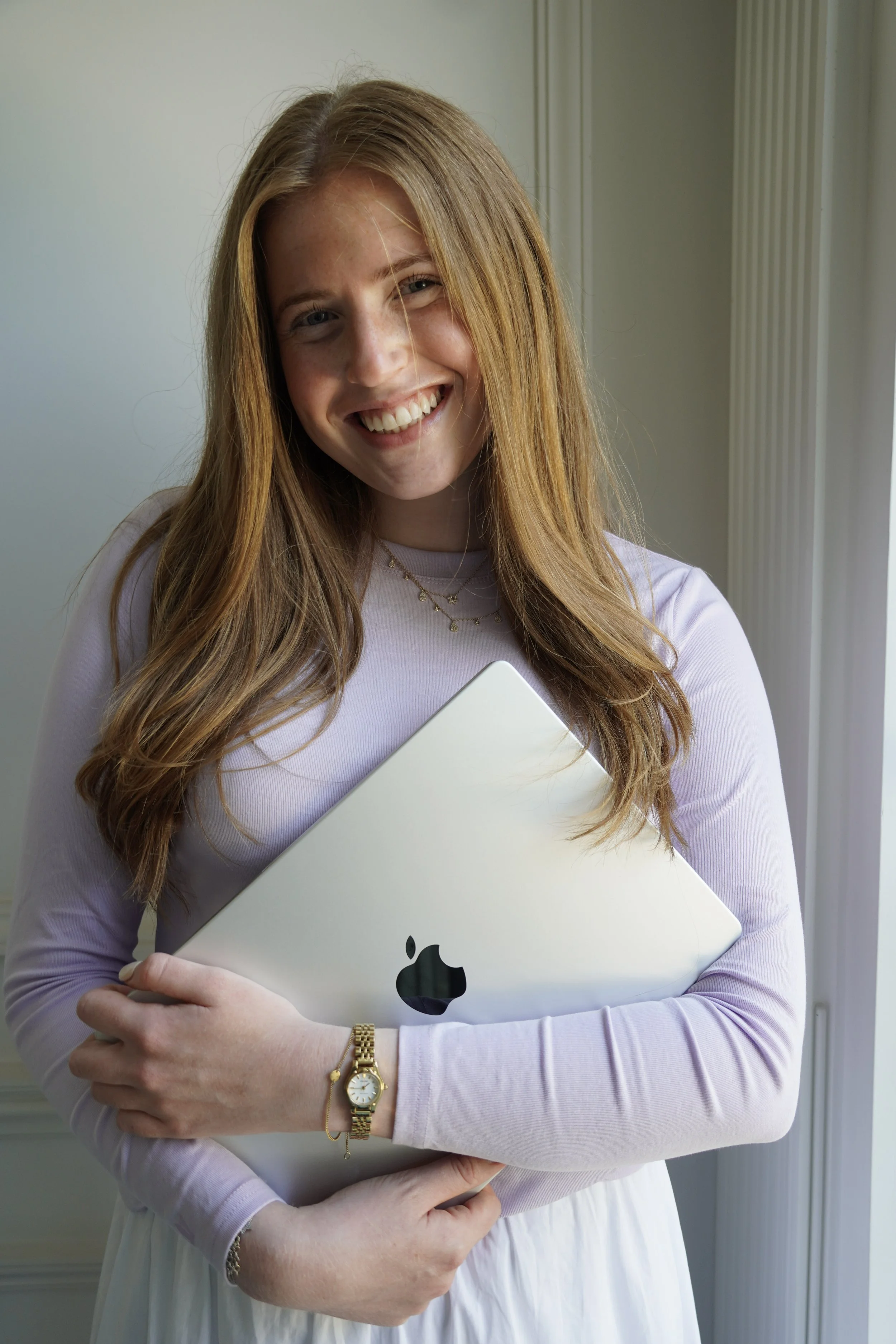 A young woman with long red hair, smiling, holding a silver MacBook laptop and wearing a pink long-sleeve shirt, a gold watch, a necklace, and a bracelet.