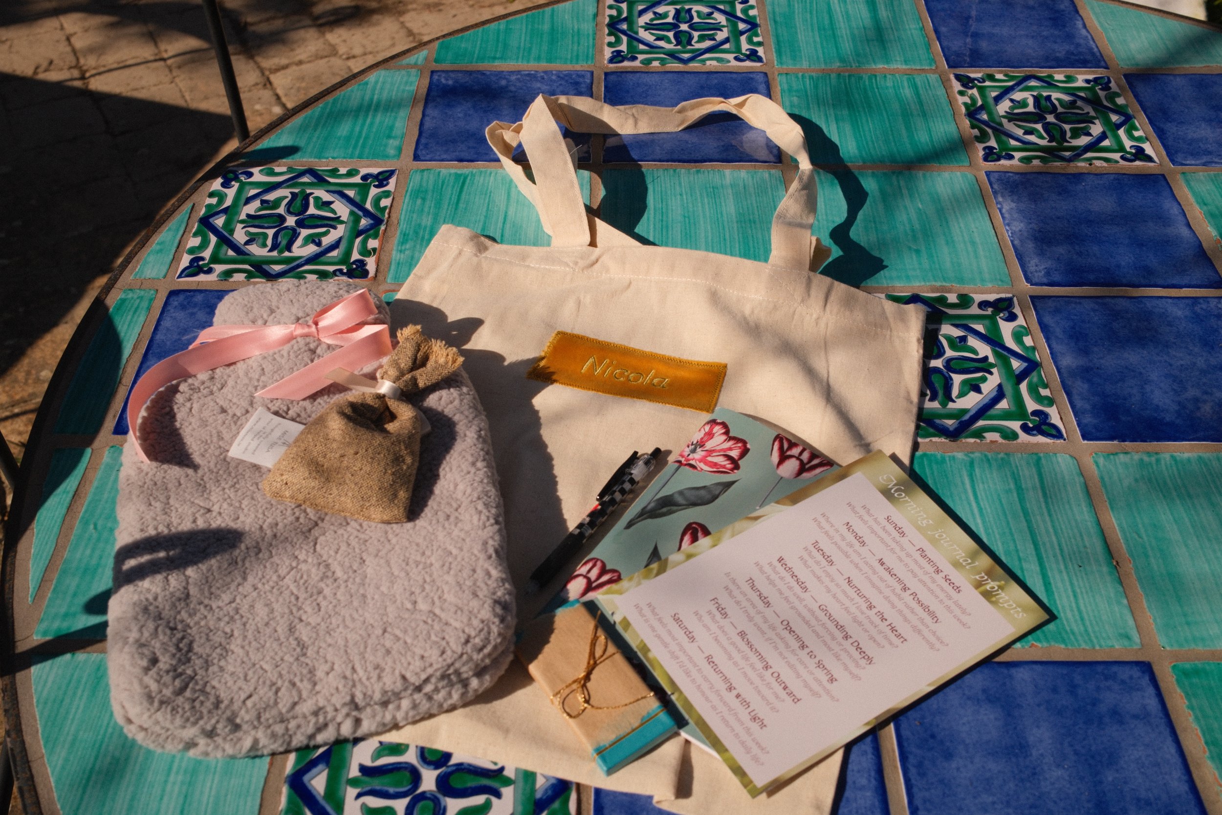 A colorful round table with a mosaic tile top featuring blue and green patterns. On the table are a white tote bag with a yellow name tag reading 'Nicola', a pink fuzzy item with a pink ribbon, a small burlap pouch, a floral booklet, and a printed sc