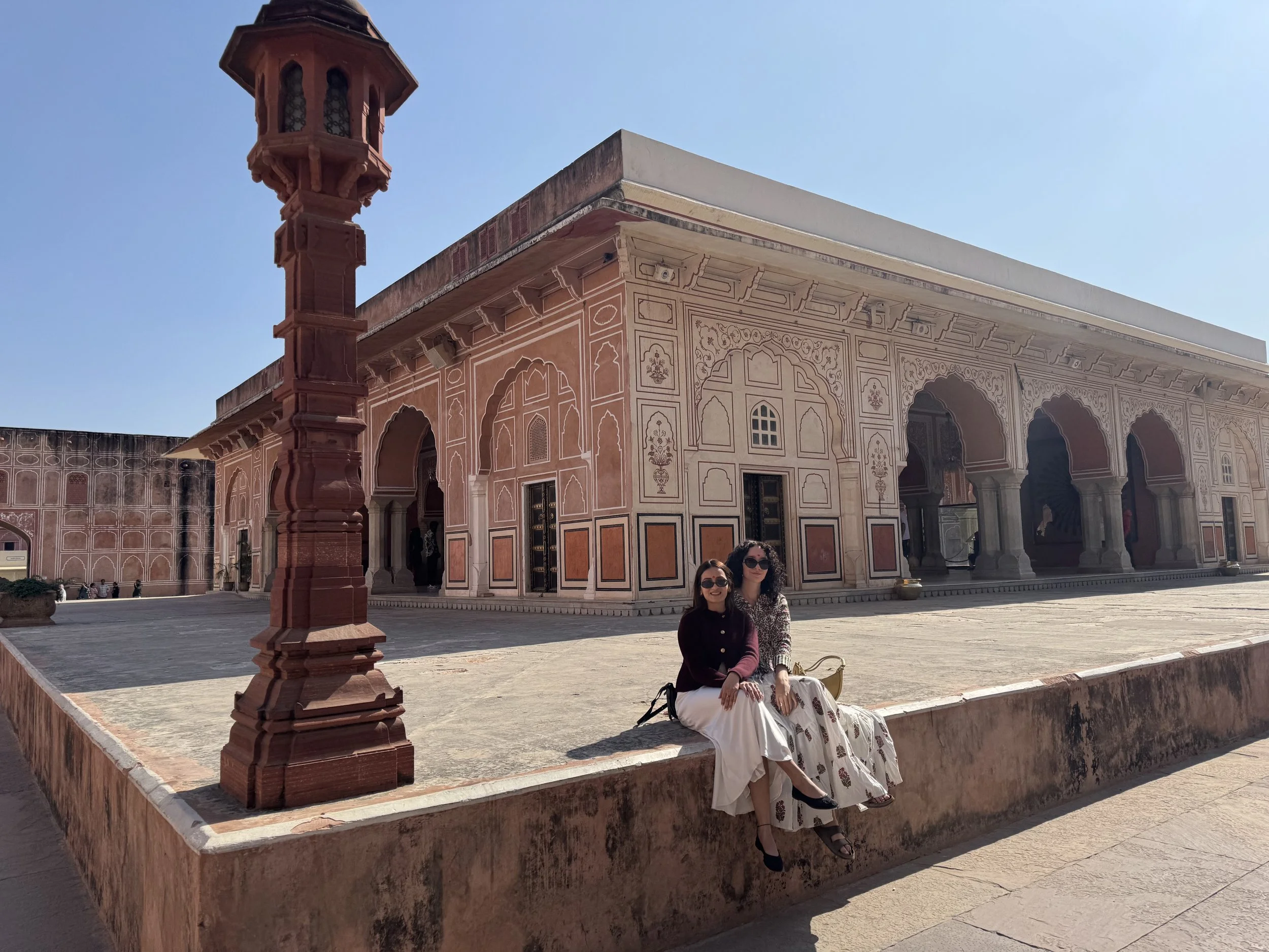 Two women sitting on a low wall in front of a historic pink sandstone building with arches and intricate carvings, under a clear blue sky.  Meala Soul India retreat jaipur yoga retreat