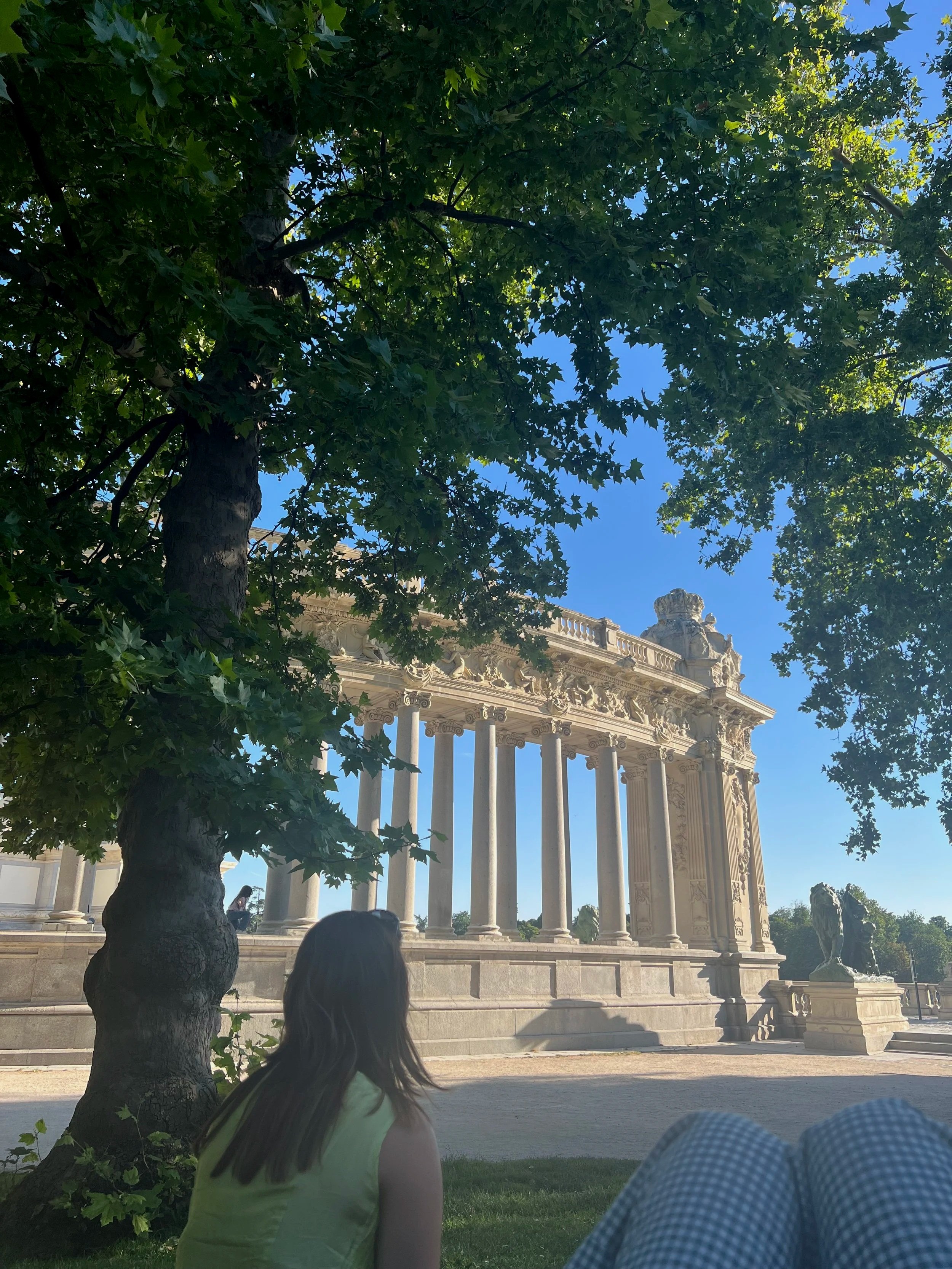 Person sitting on grass under a leafy tree, looking towards a classical architectural structure with columns and statues, under a clear blue sky. Madrid yoga retreat weekend meet-up group meala soul