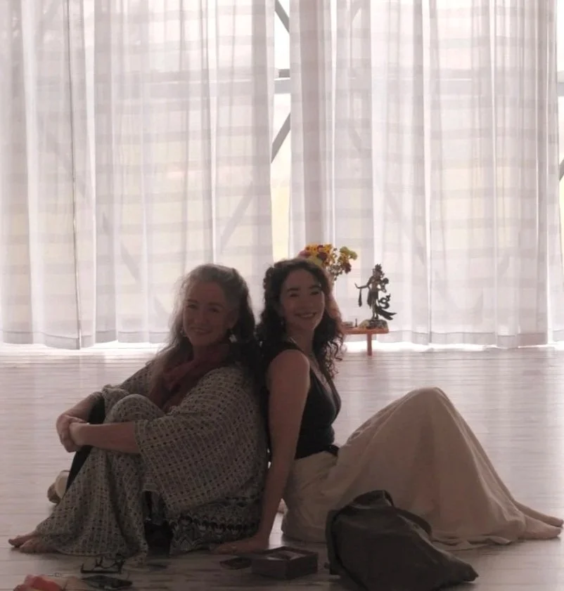 Two women sitting back to back on a wooden floor in front of white curtains, smiling. Behind them, a small table with a vase of flowers and a decorative figurine. Natural light coming through the curtains. Bridget and Meritta Meala Soul