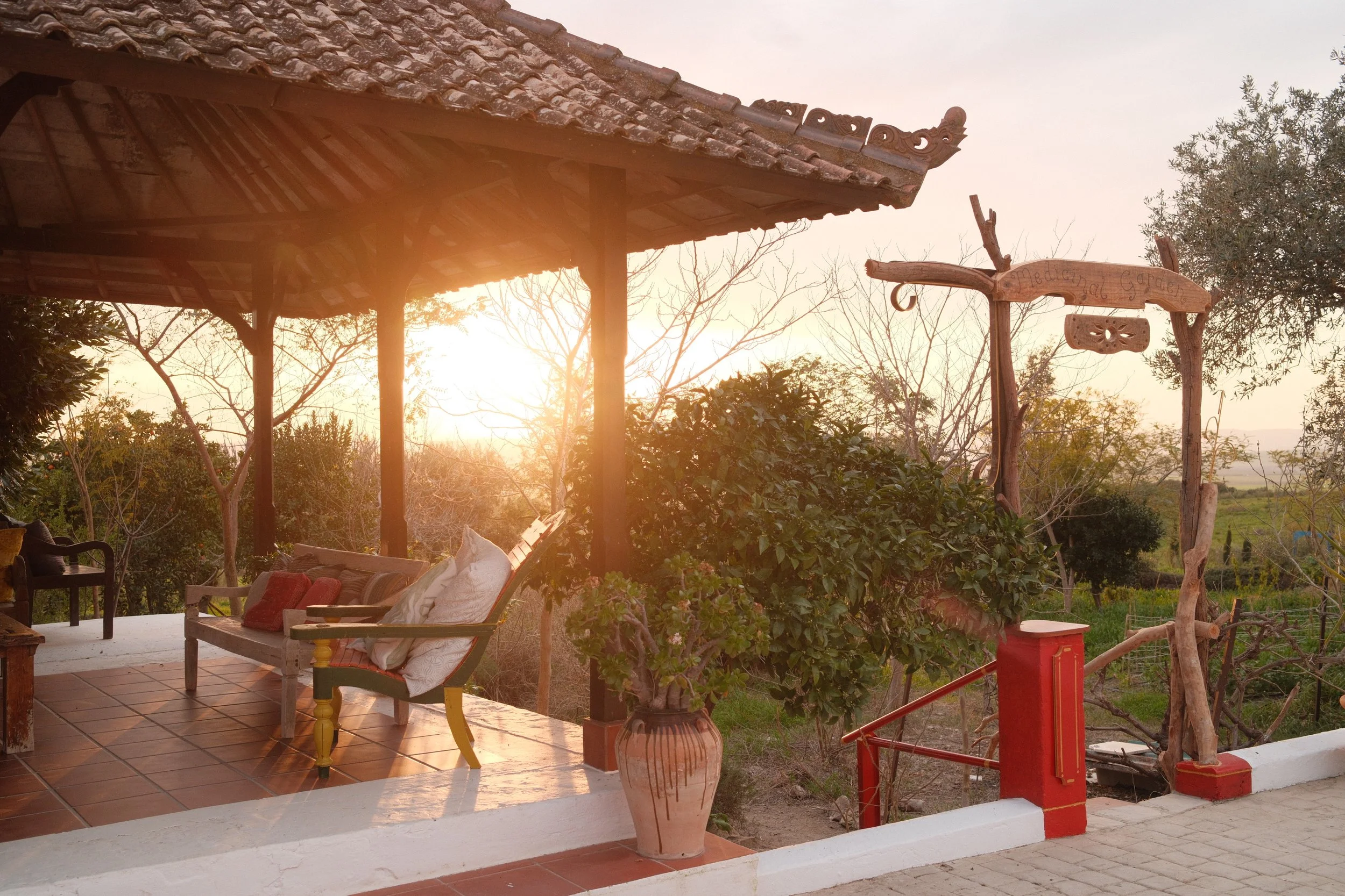 Outdoor patio area with wooden benches, pillows, potted plant, and a wooden sign, during sunset or sunrise with trees and scenic landscape in the background.