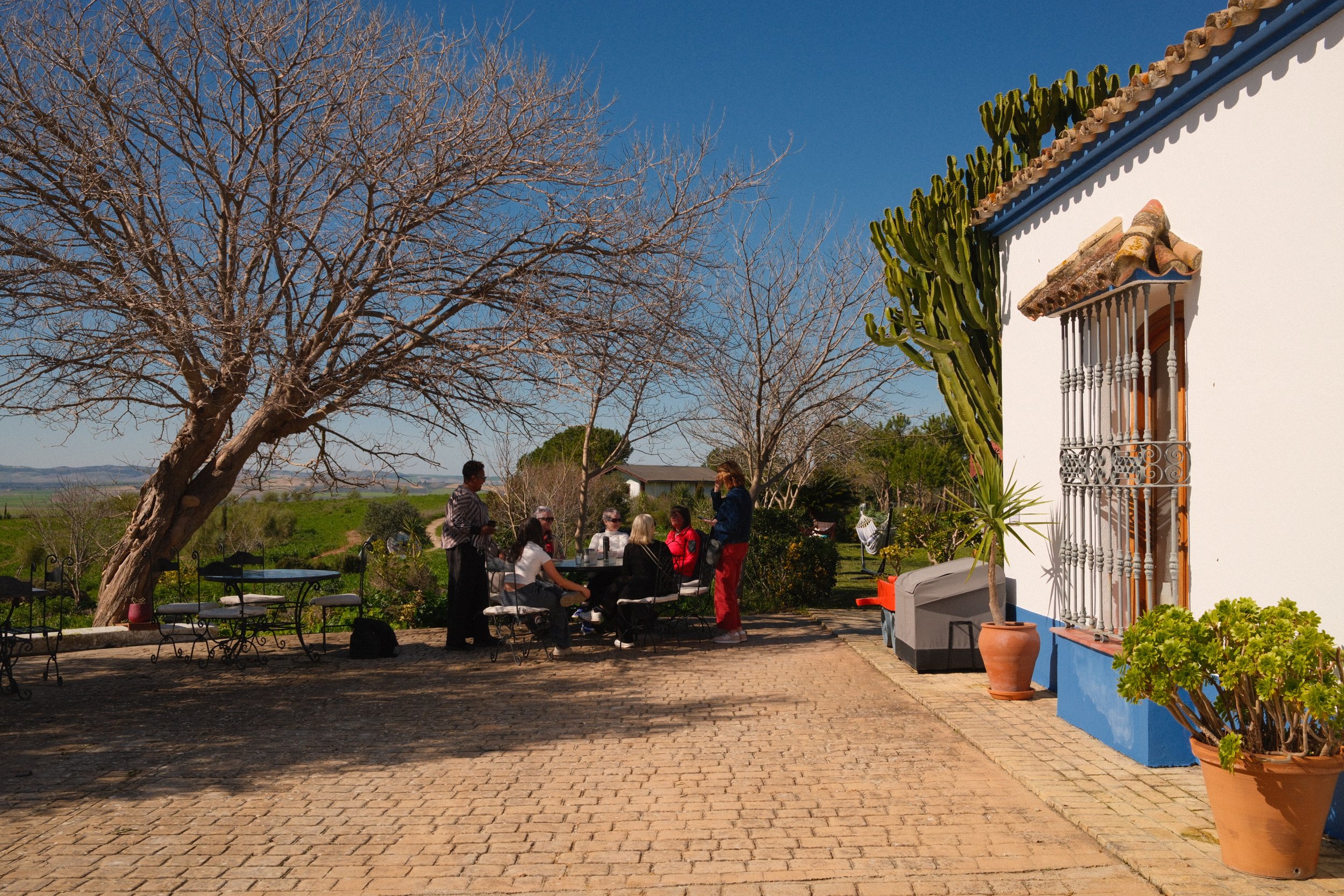 People sitting and standing around a table outdoors on a patio, with leafless trees and fields in the background, next to a white building with potted plants and a window with iron bars. Meala Soul Yoga wellness retreat spain india womens retreat