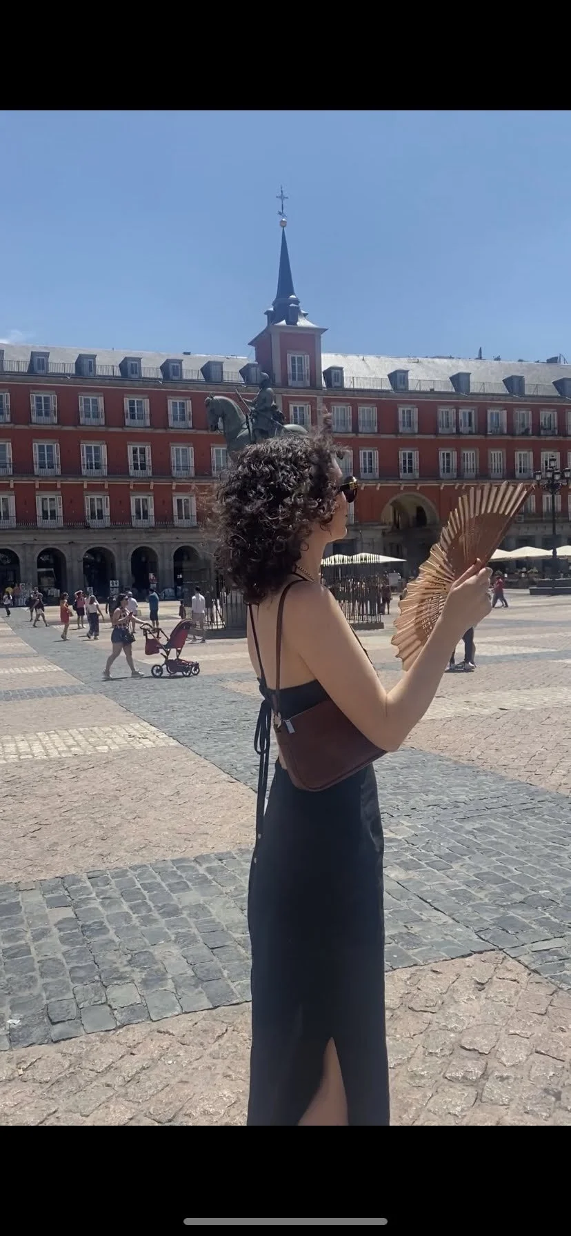 A woman with curly hair wearing a black dress and sunglasses holds a fan, standing in a European city square with historic buildings and a statue in the background.