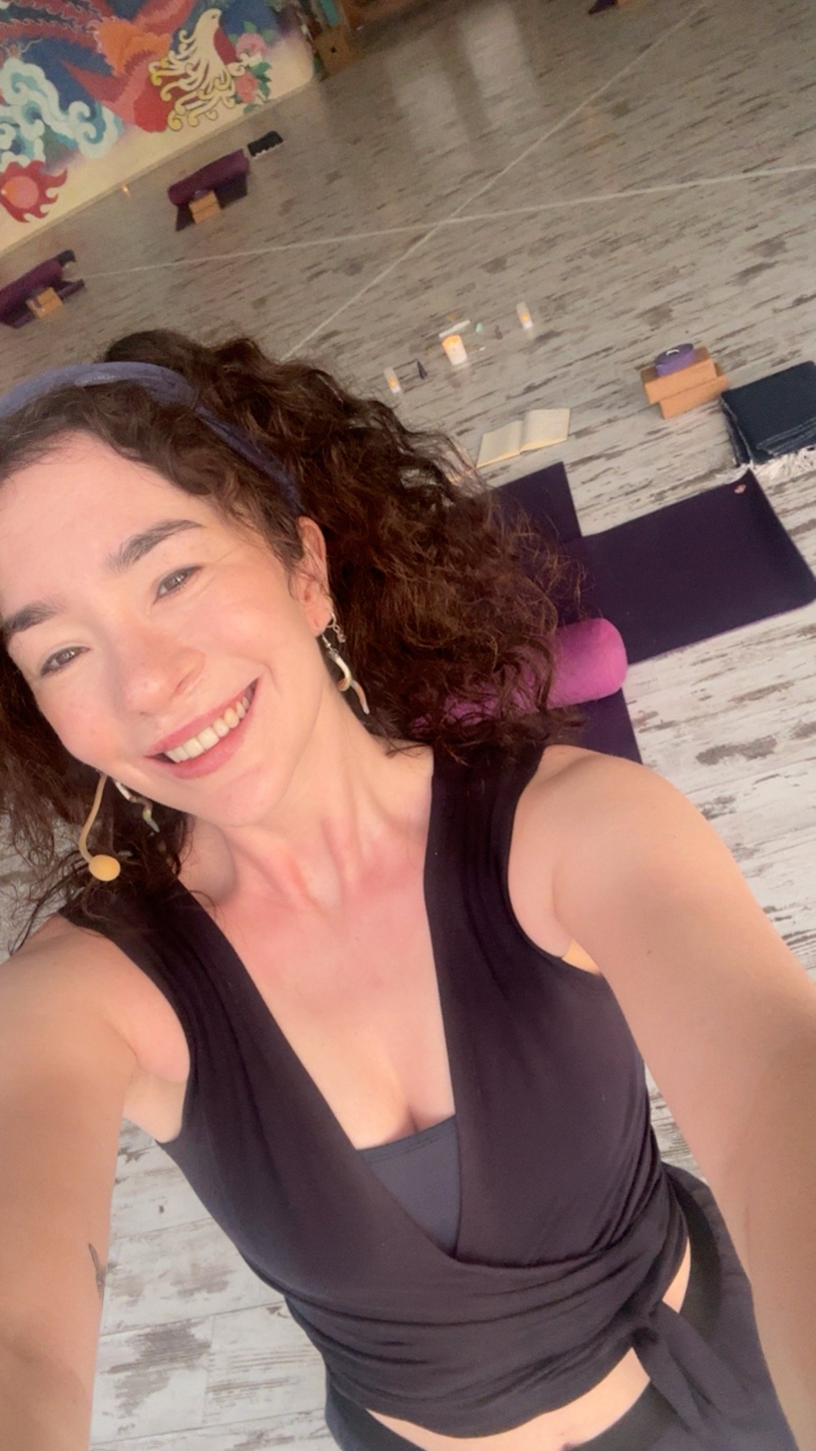 A woman with curly dark hair, smiling, lying on her back in a yoga studio with a yoga mat, meditation supplies, and colorful wall art in the background. Bridget Meala Soul yoga retreats 