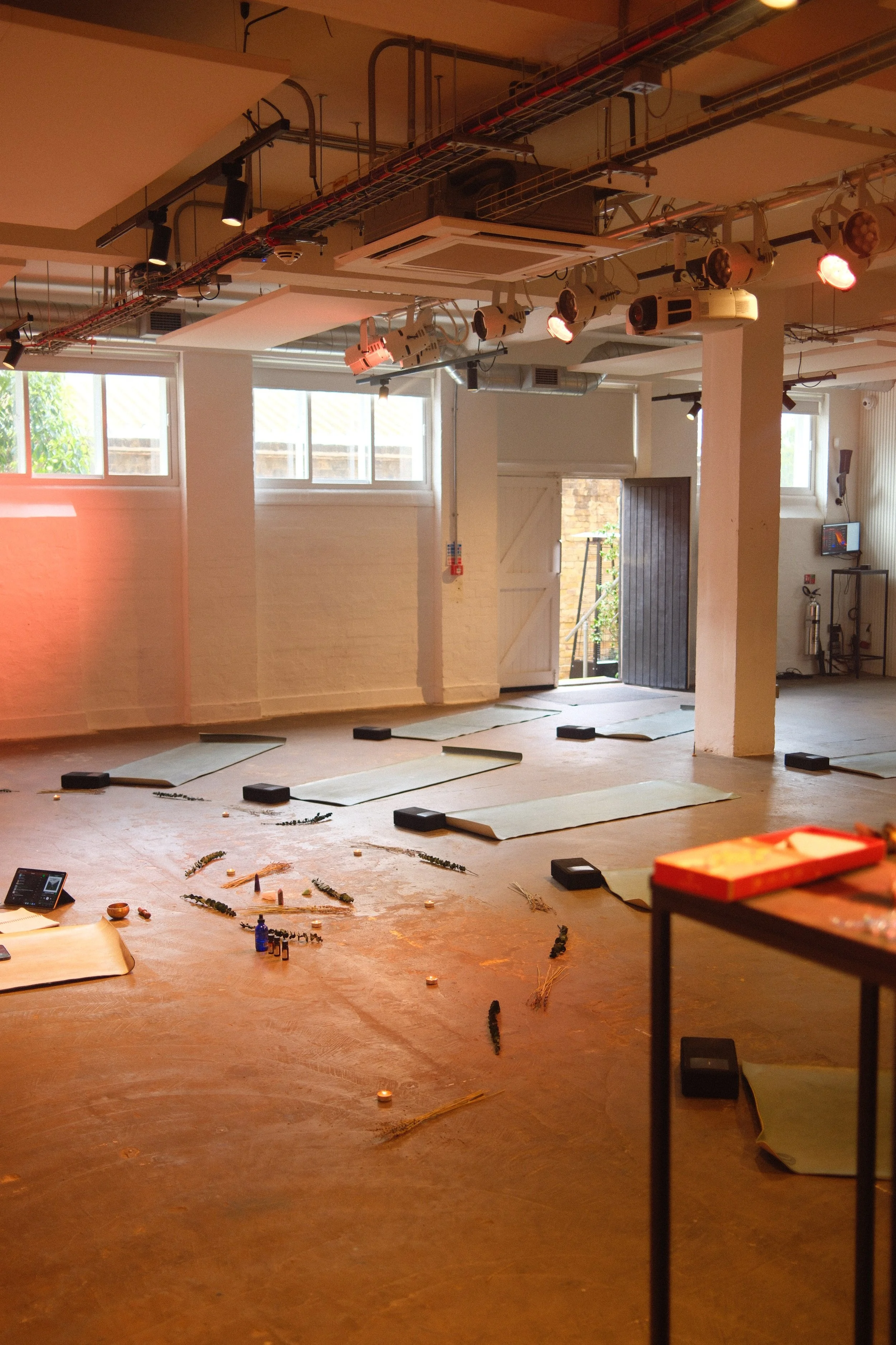 Empty indoor space with yoga mats and scattered objects on the floor, with some lighting equipment on the ceiling and an open door leading outside.