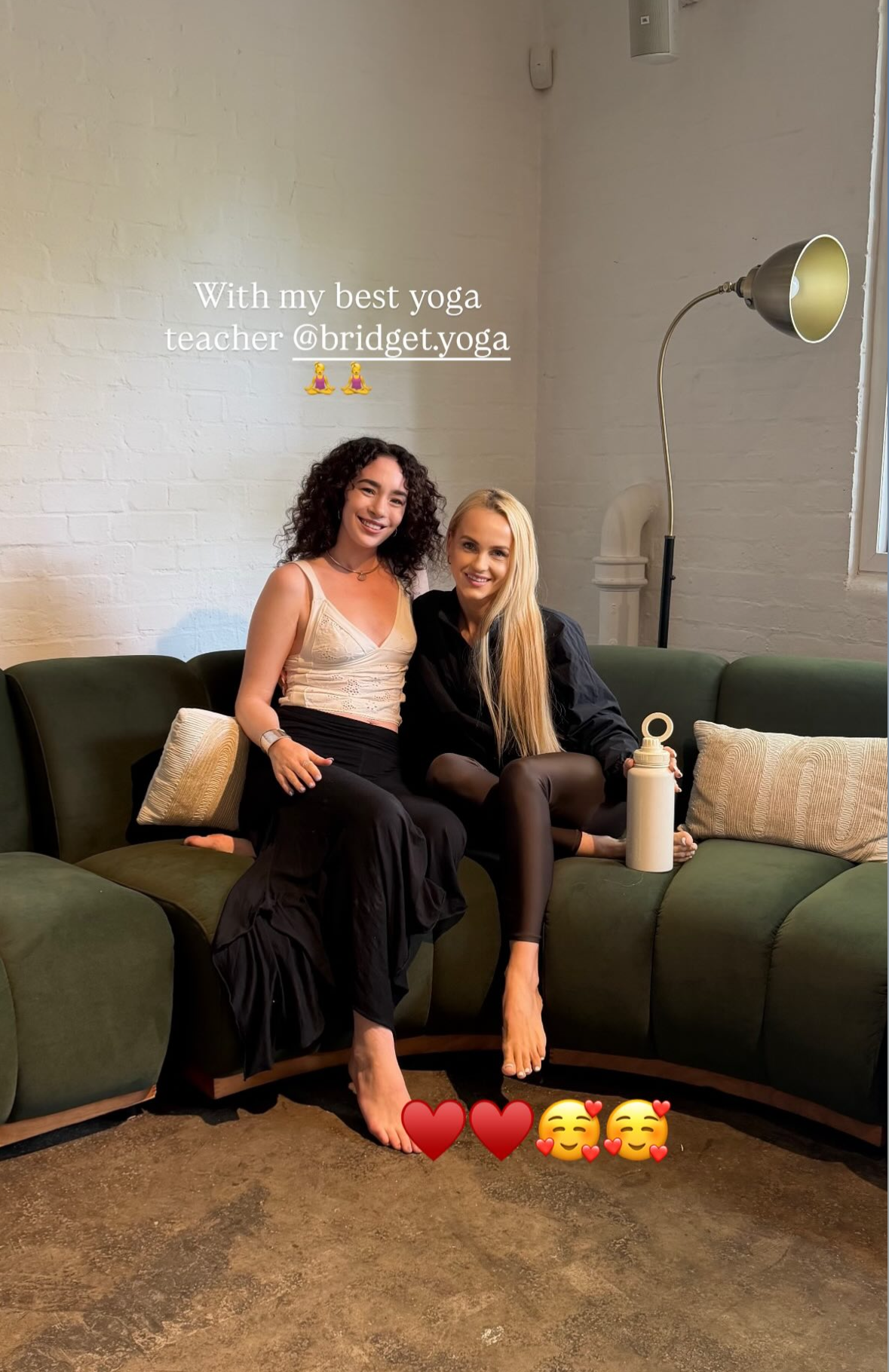 Two women sitting on a dark green couch, smiling. One has curly dark hair and is wearing a beige top and black pants, the other has long blonde hair and is wearing a black jacket and black tights. There are pillows on the couch, and a white water bot