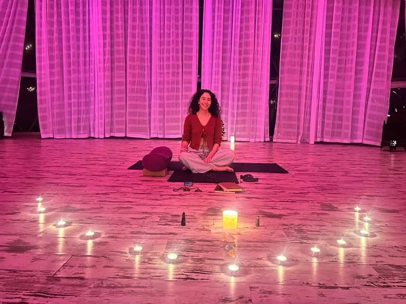 Woman sitting cross-legged on yoga mat in front of pink curtains, surrounded by candles, brought yoga props, and a small altar, in a cozy indoor space. Meala Soul Yoga Nidra