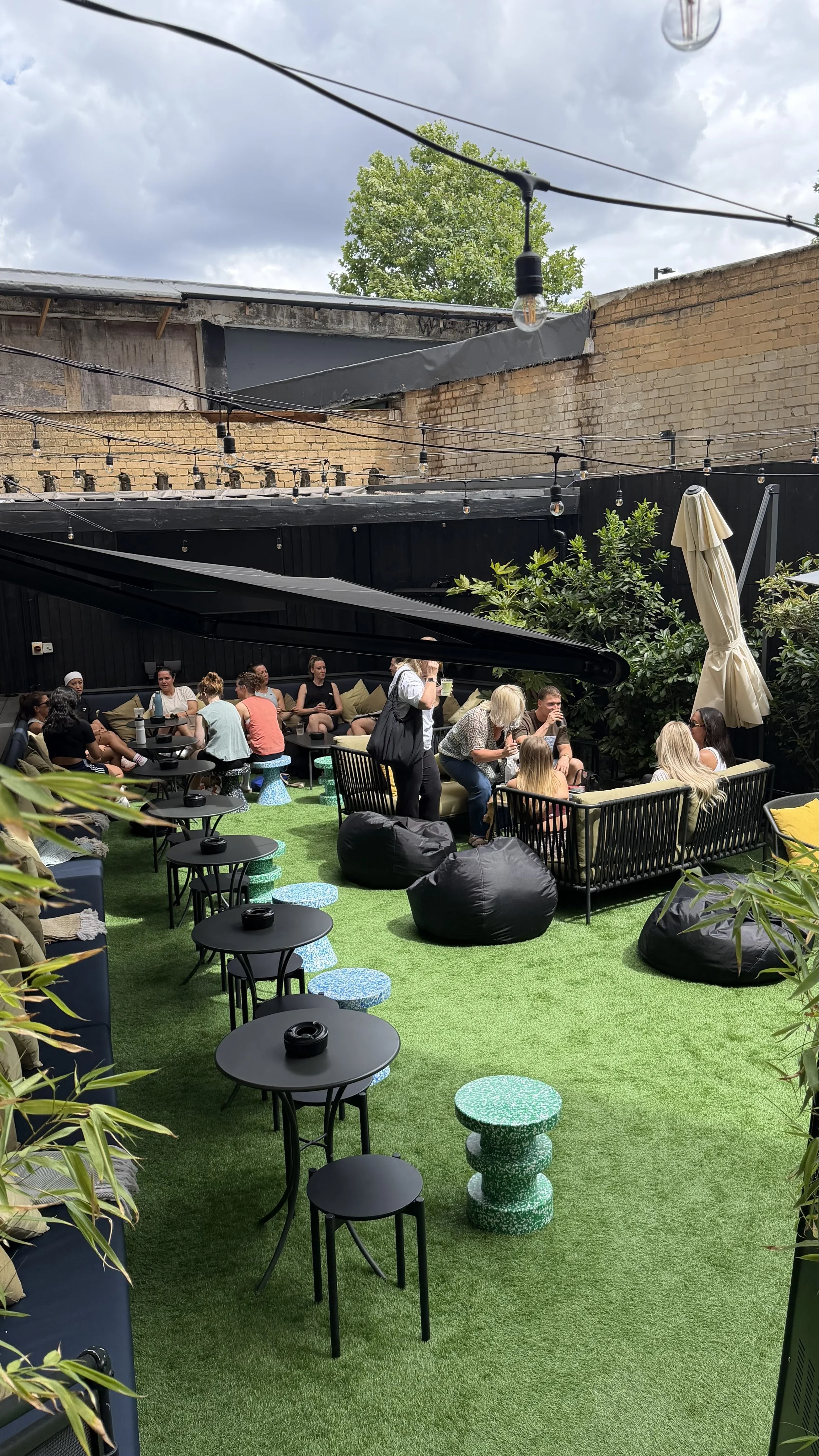 People sitting and socializing in an outdoor patio with string lights, black tables, and green artificial turf, surrounded by brick and black painted walls, with trees and cloudy sky overhead.