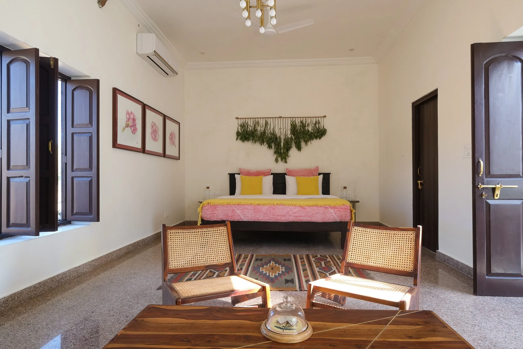 Hotel room with a bed, wall art, wooden chairs, a table with a glass dome, open window, and indoor plants.  Meala Soul India retreat jaipur yoga retreat