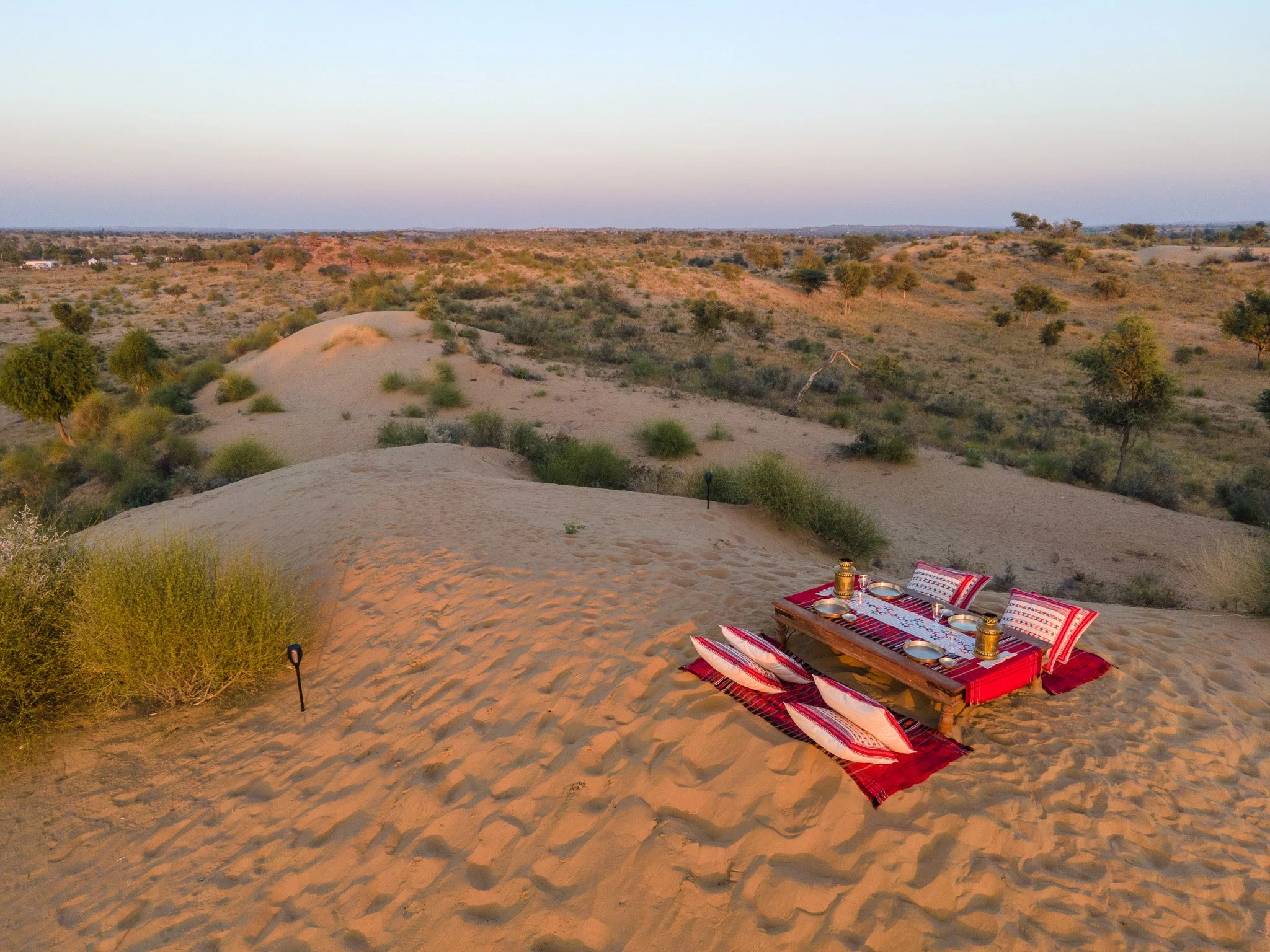 Desert landscape with scattered bushes and small trees, a sunset sky, and a low table set for outdoor dining with cushions, pillows, and lanterns on sand..  Meala Soul India retreat jaipur yoga retreat