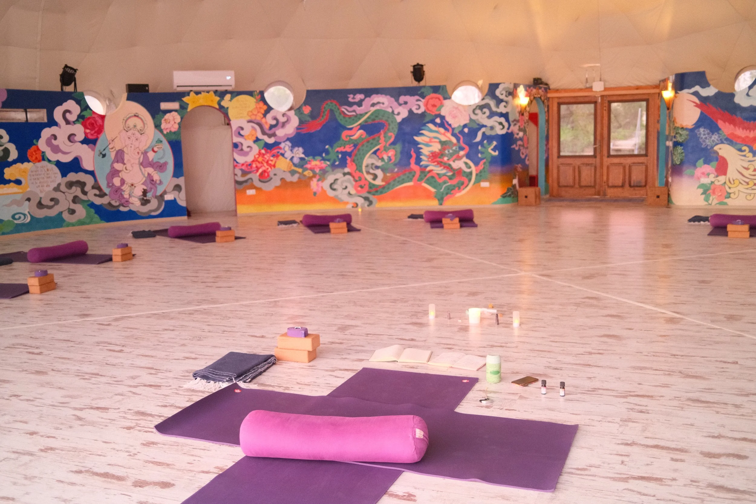A spacious yoga studio with a colorful mural of spiritual figures and mythical creatures on the walls, floor mats, yoga props, and candles arranged for practice.