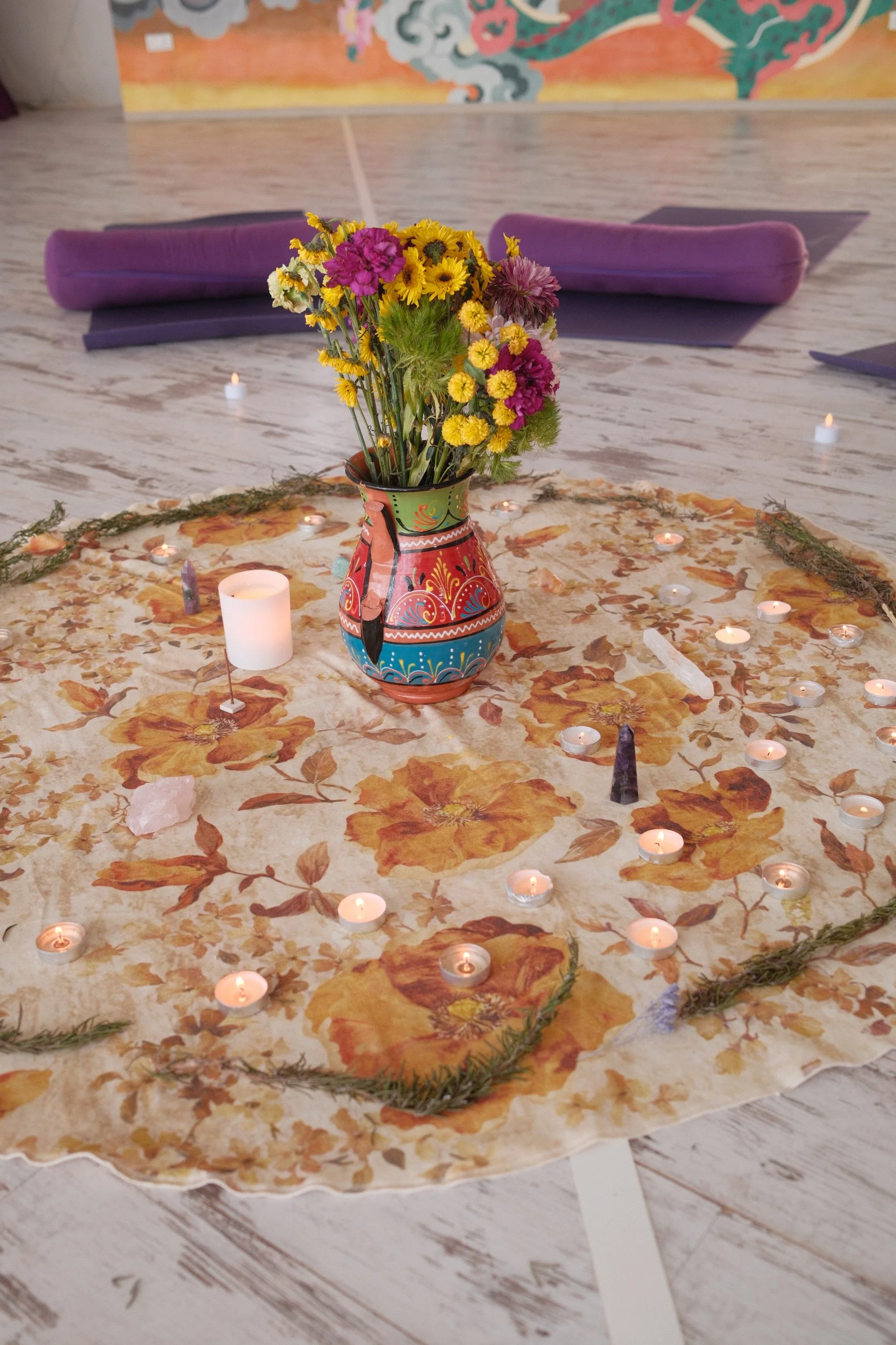 A floral arrangement with yellow, purple, and white flowers in a decorative vase placed on a fabric mat with orange and yellow floral patterns. Surrounding the arrangement are small tea light candles, candles, and pauses, with purple yoga mats and a 