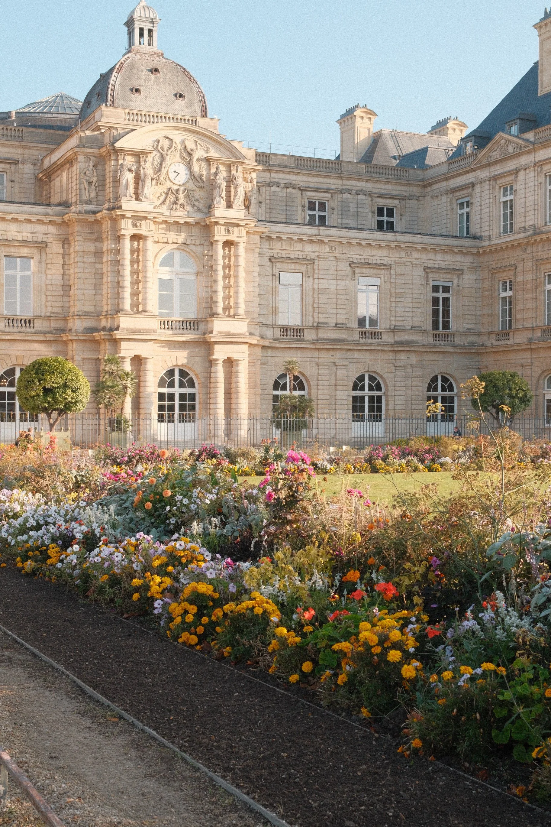 A historic beige stone building with ornate classical architecture, a domed roof, and a clock. In front, a garden with colorful flowers and trimmed bushes. Bridget Meala Soul yoga retreats 