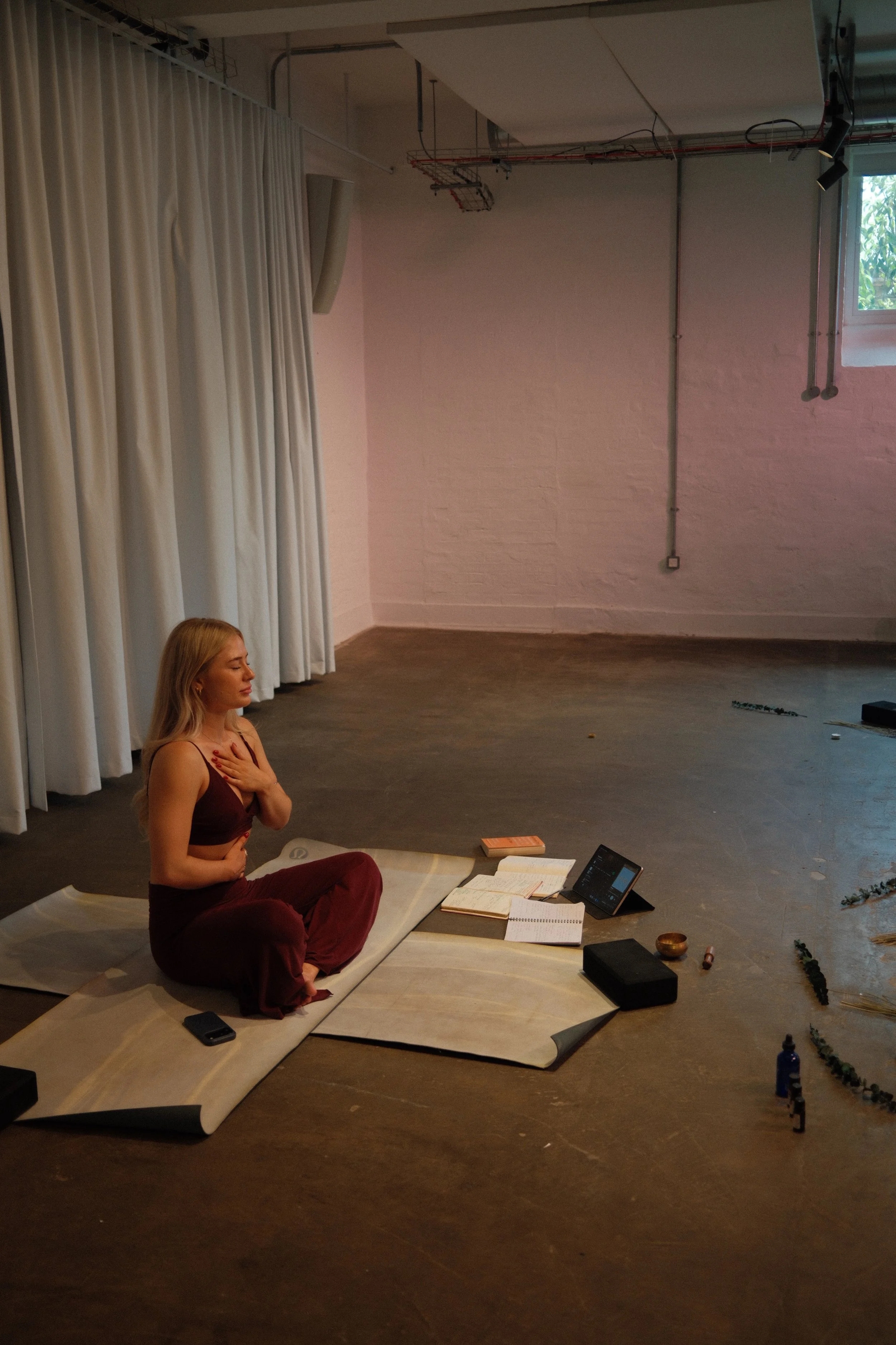 A woman practicing meditation or yoga in a peaceful indoor space, sitting on a mat with her eyes closed and hands over her heart. There are closed books, a tablet, and personal items around her, with a stage curtain and a small window visible in the 