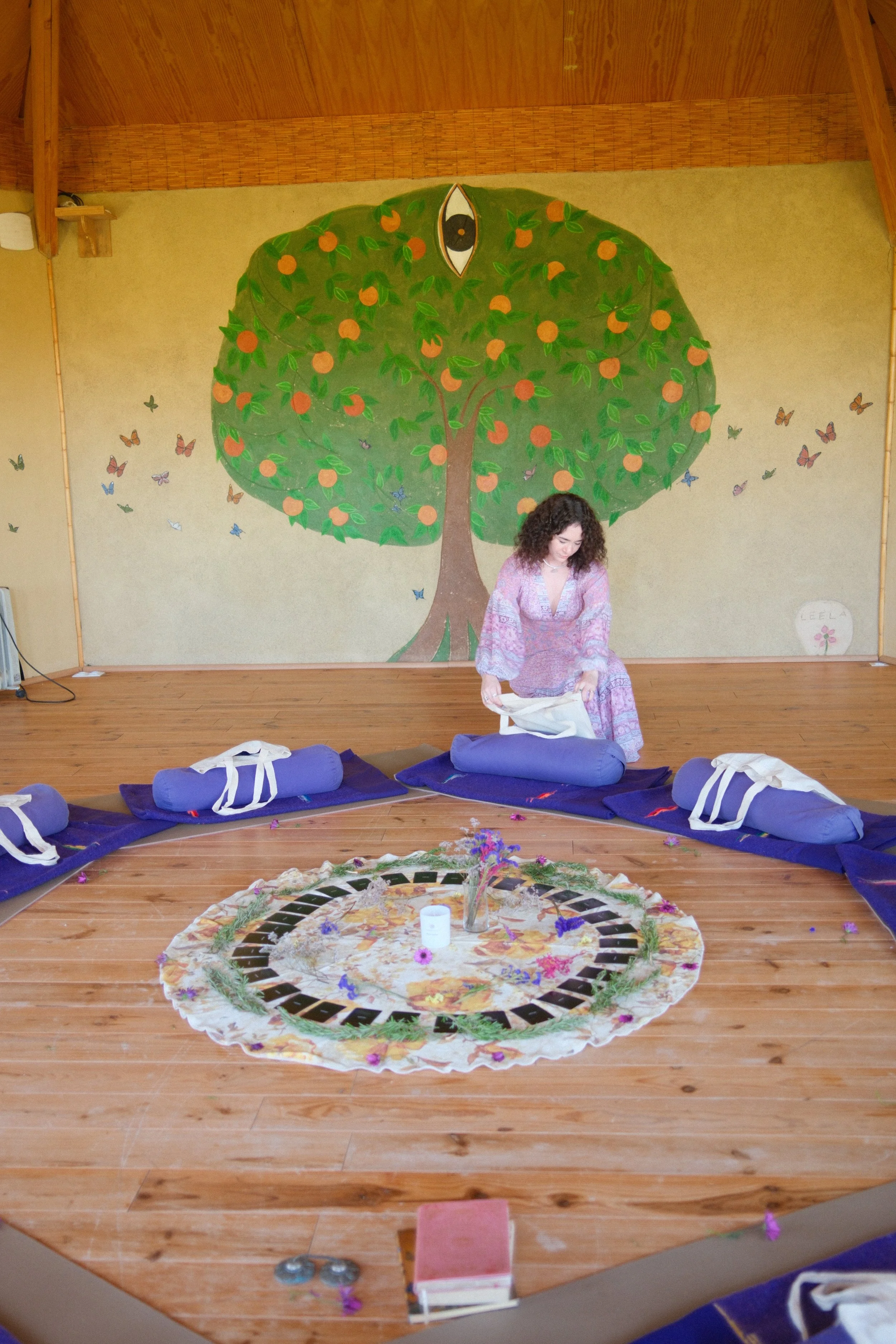 Woman in a colorful dress setting up a circle of purple mats with rolled-up blankets around a decorated cloth on a wooden floor, with a mural of a green tree with orange fruits and an eye at the top in the background.