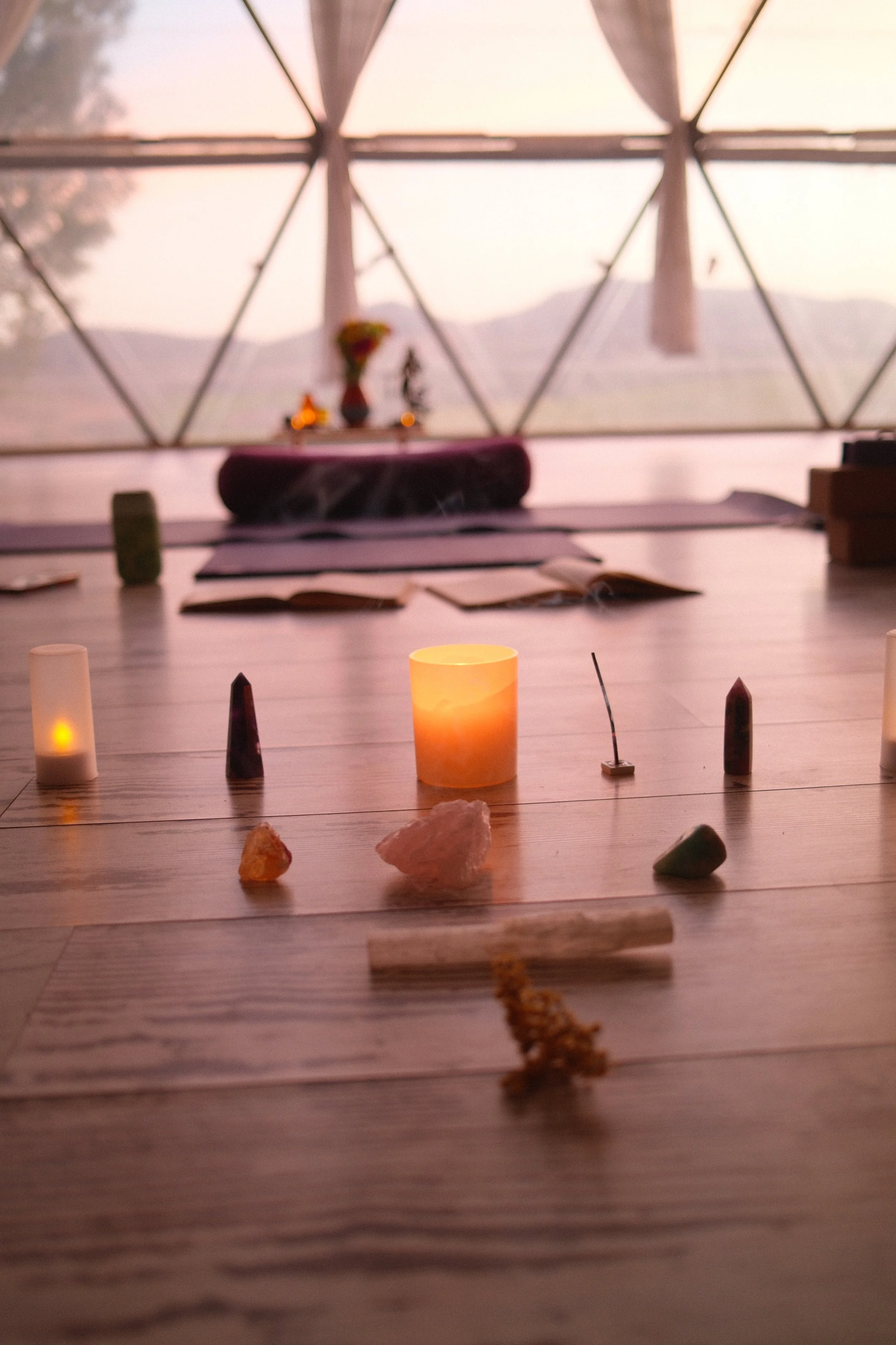 A spiritual setup inside a geodesic dome with candles and crystals on a wooden floor, overlooking mountains at sunset.